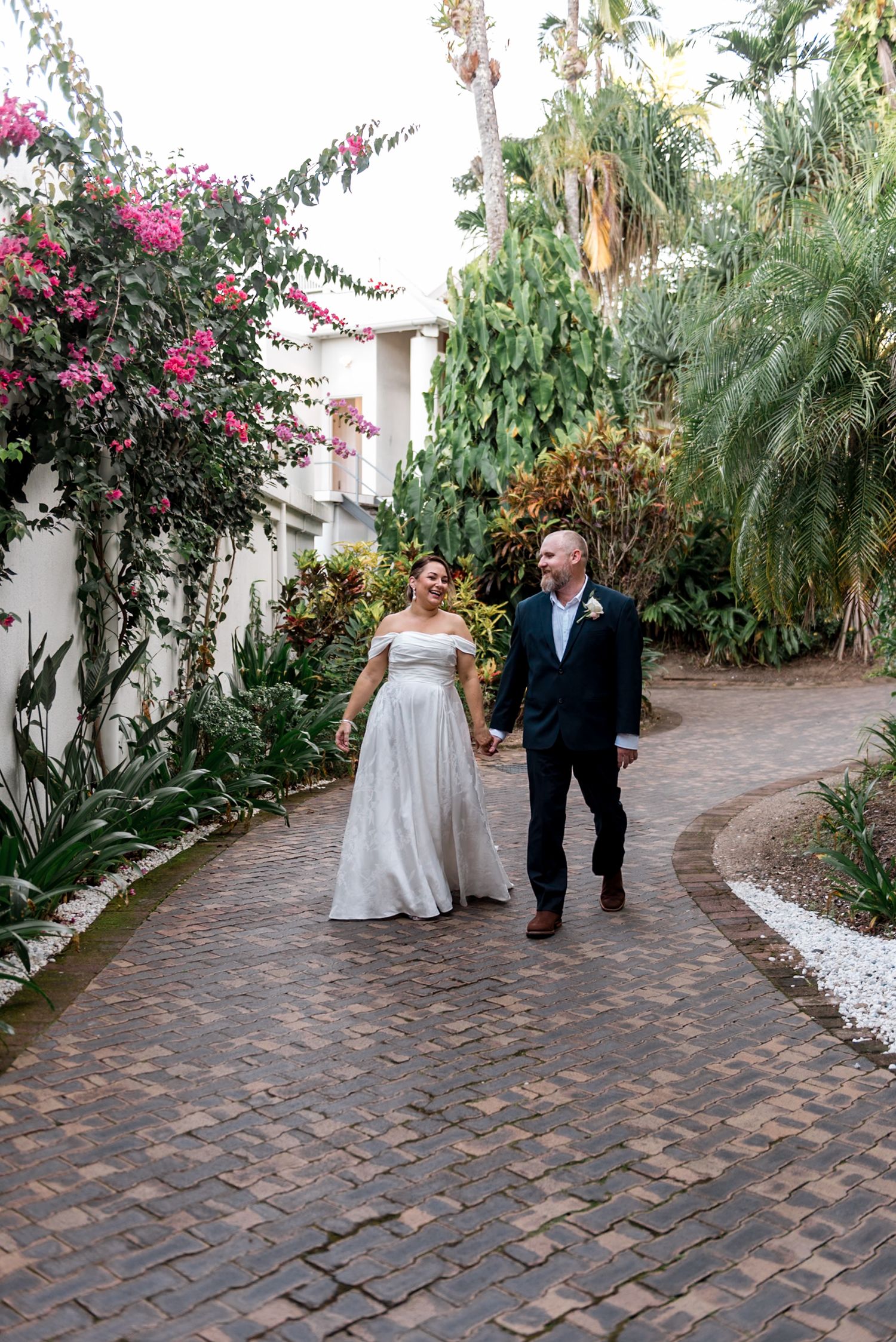 Laughing bride and groom strolling hand in hand along winding brick path in tropical garden.