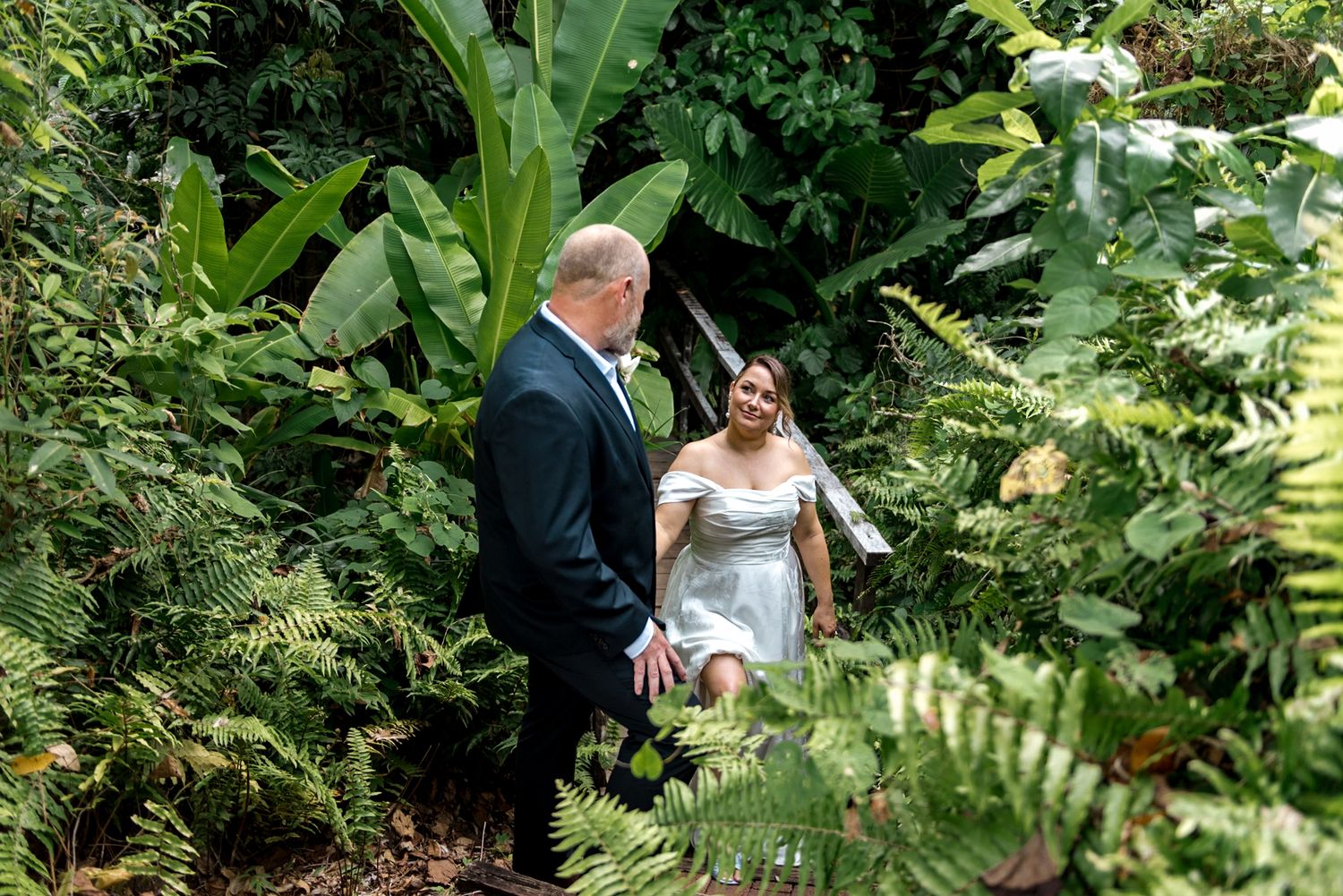 Bride in light blue wedding dress and groom in dark suit sharing romantic moment in tropical garden.