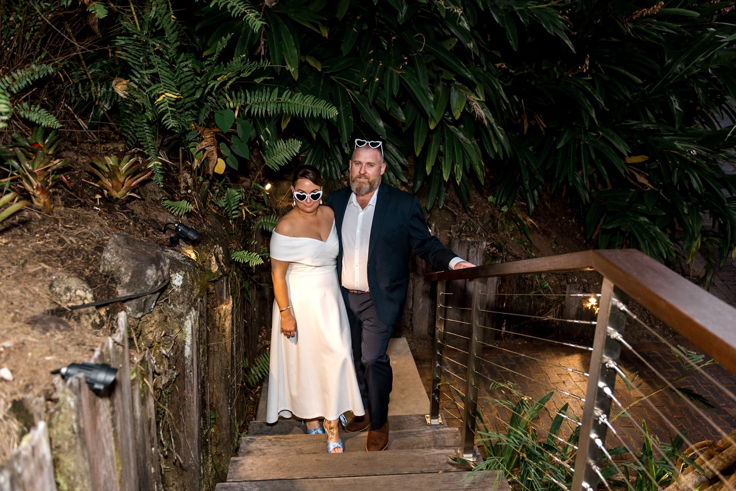 Bride in white off-shoulder dress and groom in navy suit walking up outdoor steps at night.