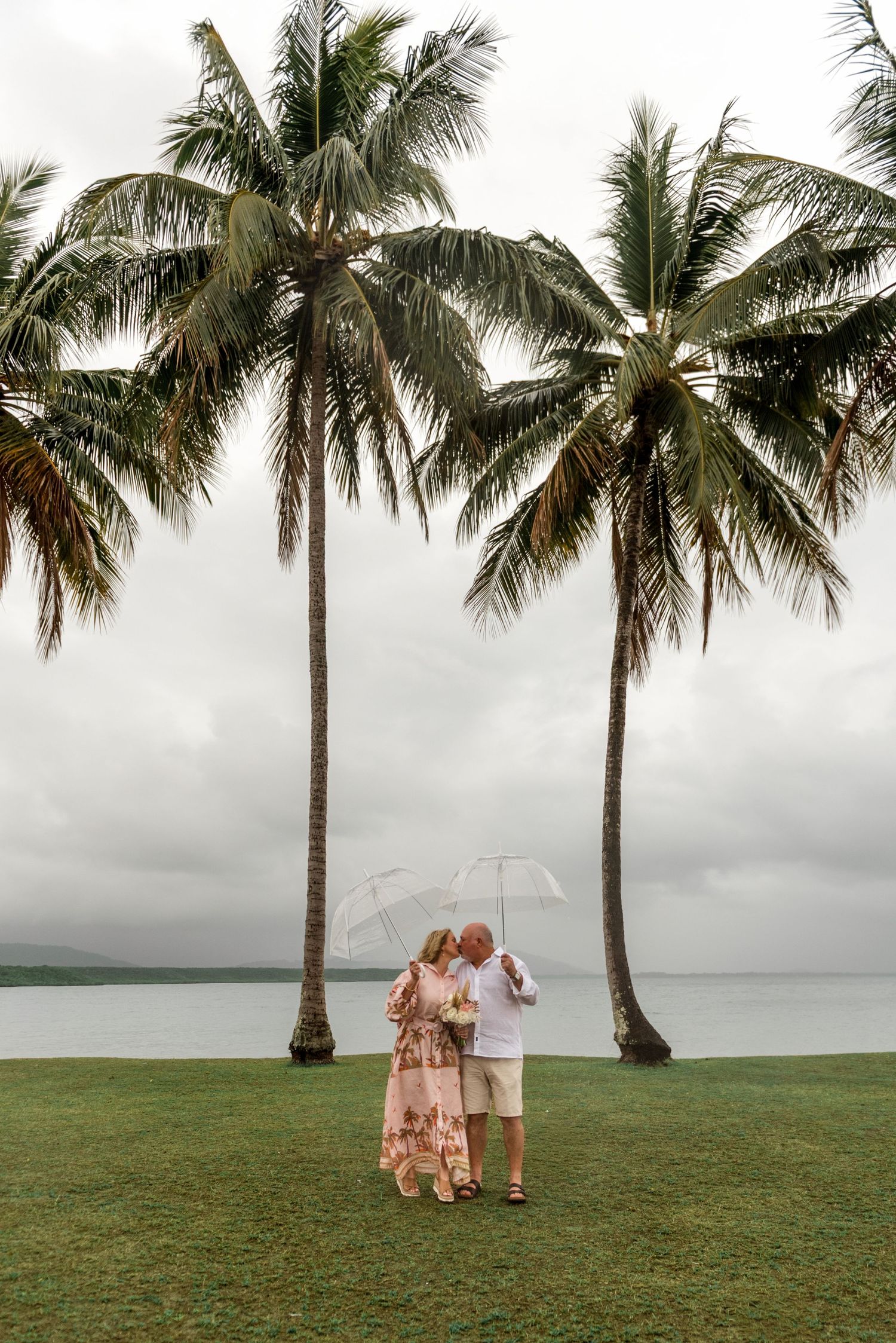 Couple kissing under a clear bubble umbrella framed by two towering palm trees on a cloudy day.