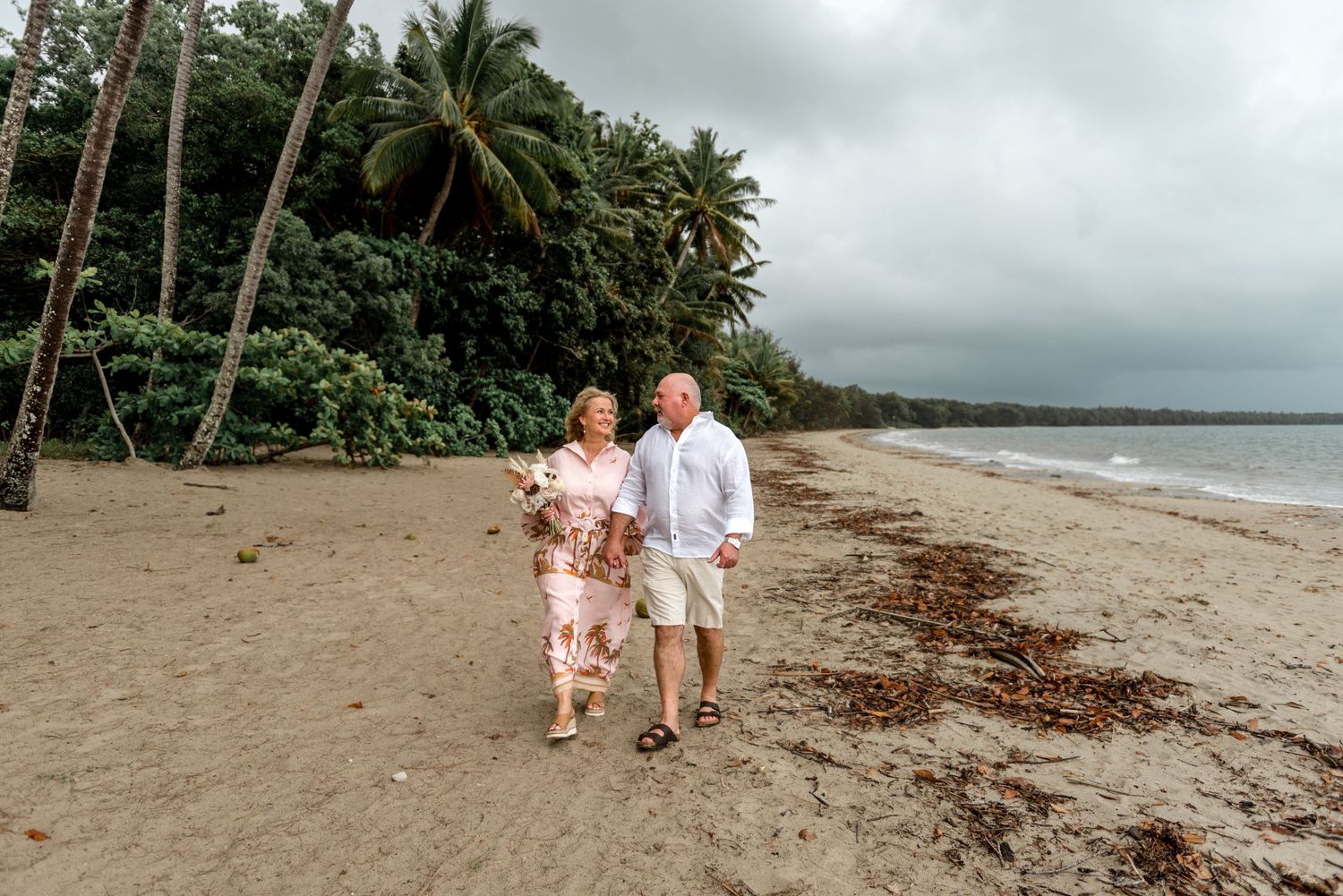 Happy mature couple walking hand in hand along a sandy tropical beach lined with palm trees.