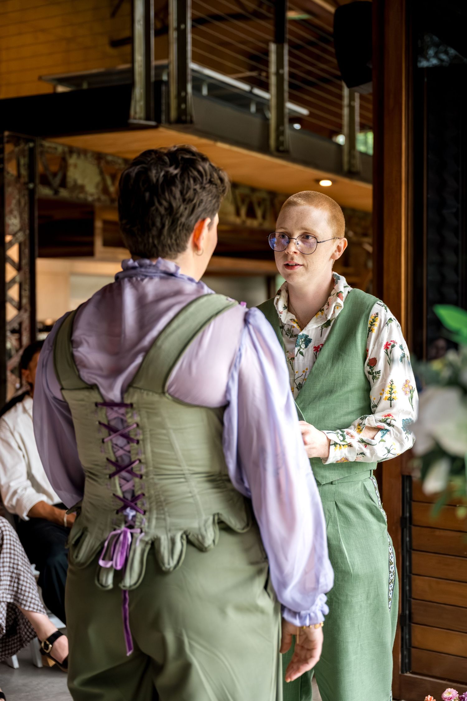 Two brides hold hands during wedding vows in green and purple Renaissance-inspired outfits.