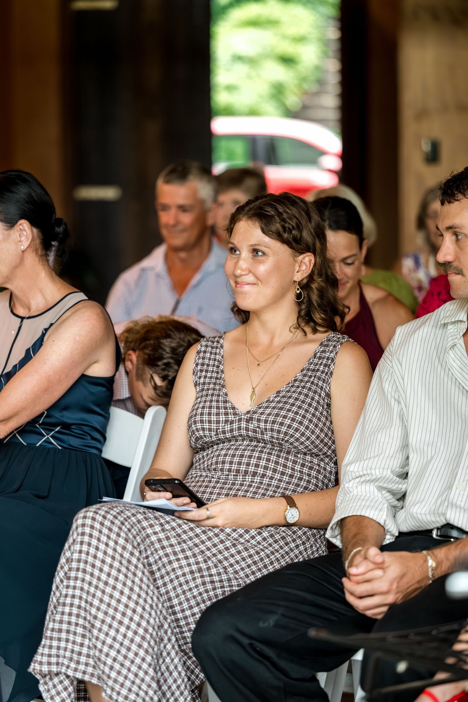 Smiling female wedding guest in checkered dress seated among other guests during wedding ceremony.