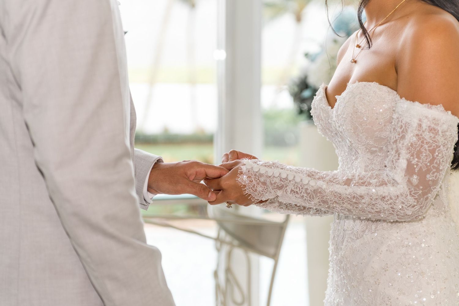 Close-up of bride in beaded lace gown and groom in gray suit holding hands at altar.