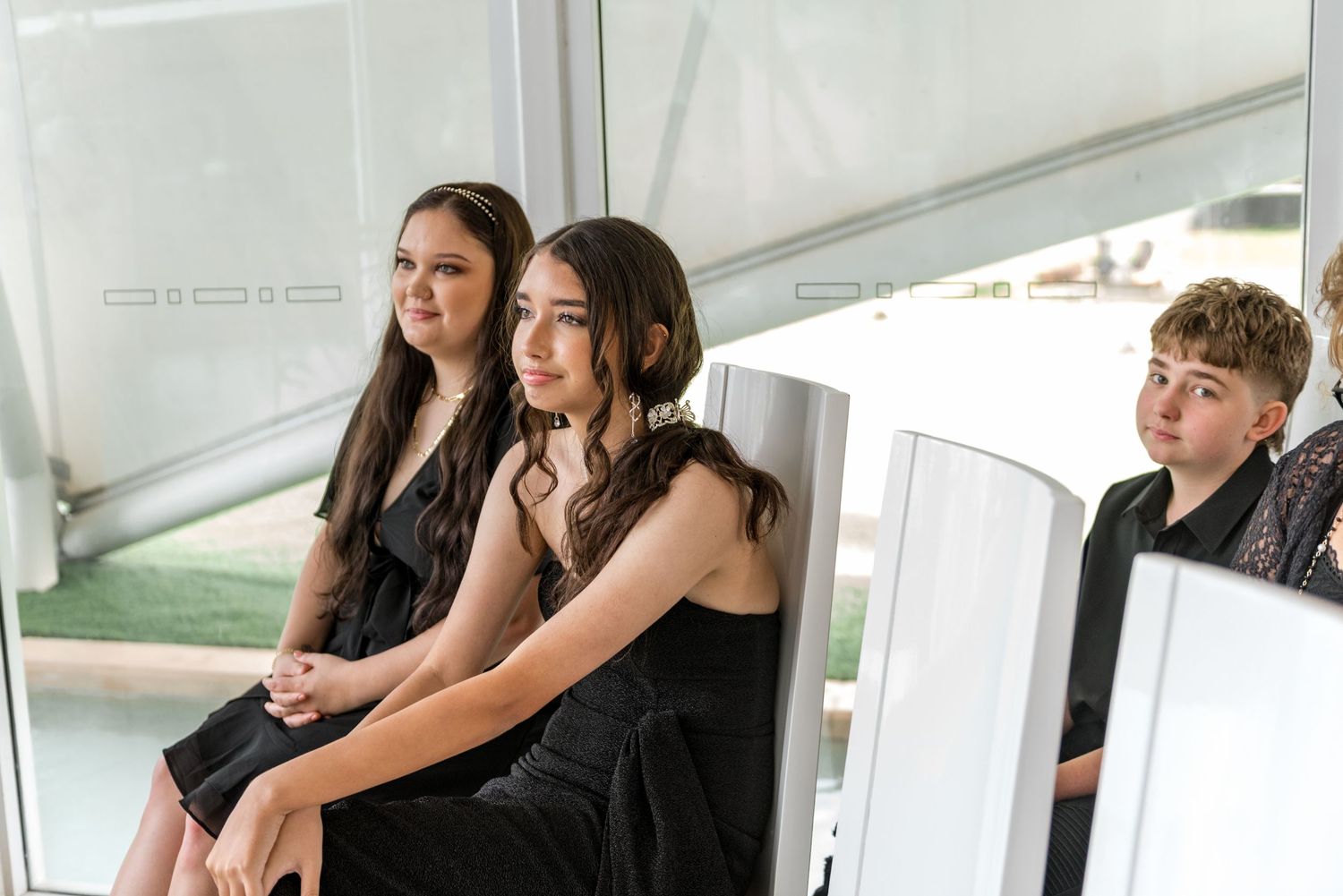 Three wedding guests in black formal attire seated together at an outdoor wedding venue.