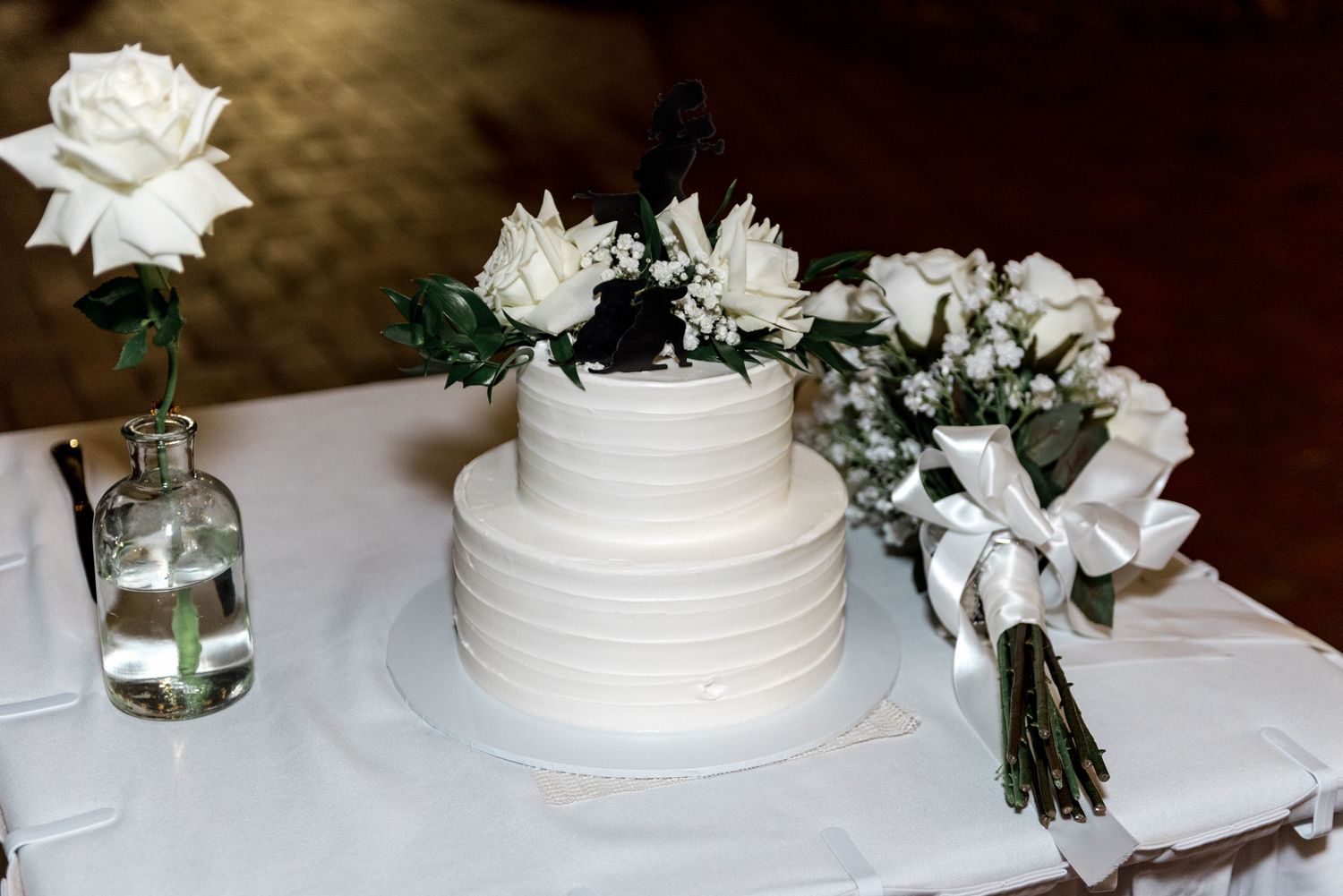 Elegant two-tier white wedding cake adorned with white roses and greenery displayed beside a bridal bouquet.