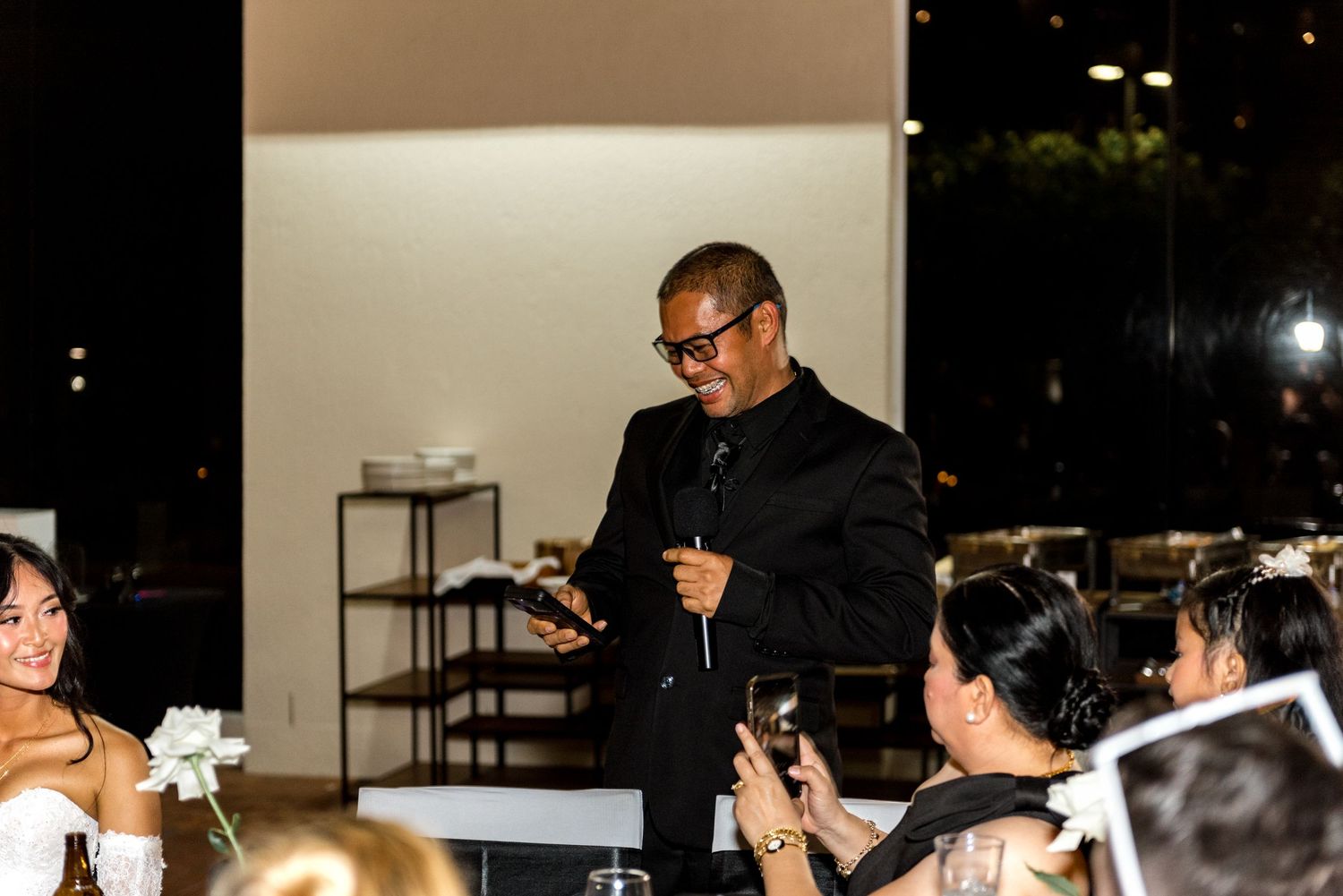 A man in a black suit gives a speech at an elegant wedding reception dinner.