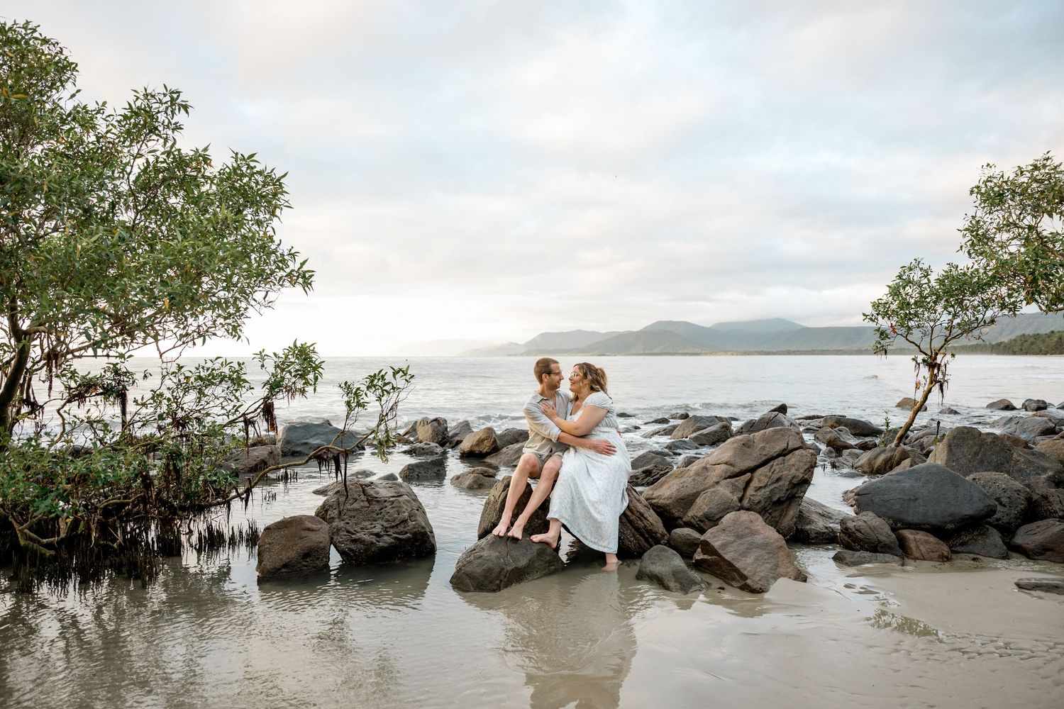 Couple sits closely on rocky shoreline surrounded by mangroves with serene ocean and mountains beyond at Four Mile Beach, Port Douglas.