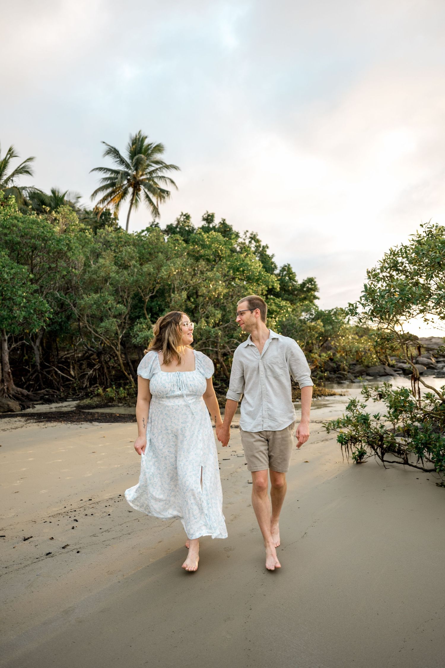 Happy couple walks hand-in-hand along tropical beach at sunset, woman in white dress, lush palms behind at Four Mile Beach, Port Douglas.