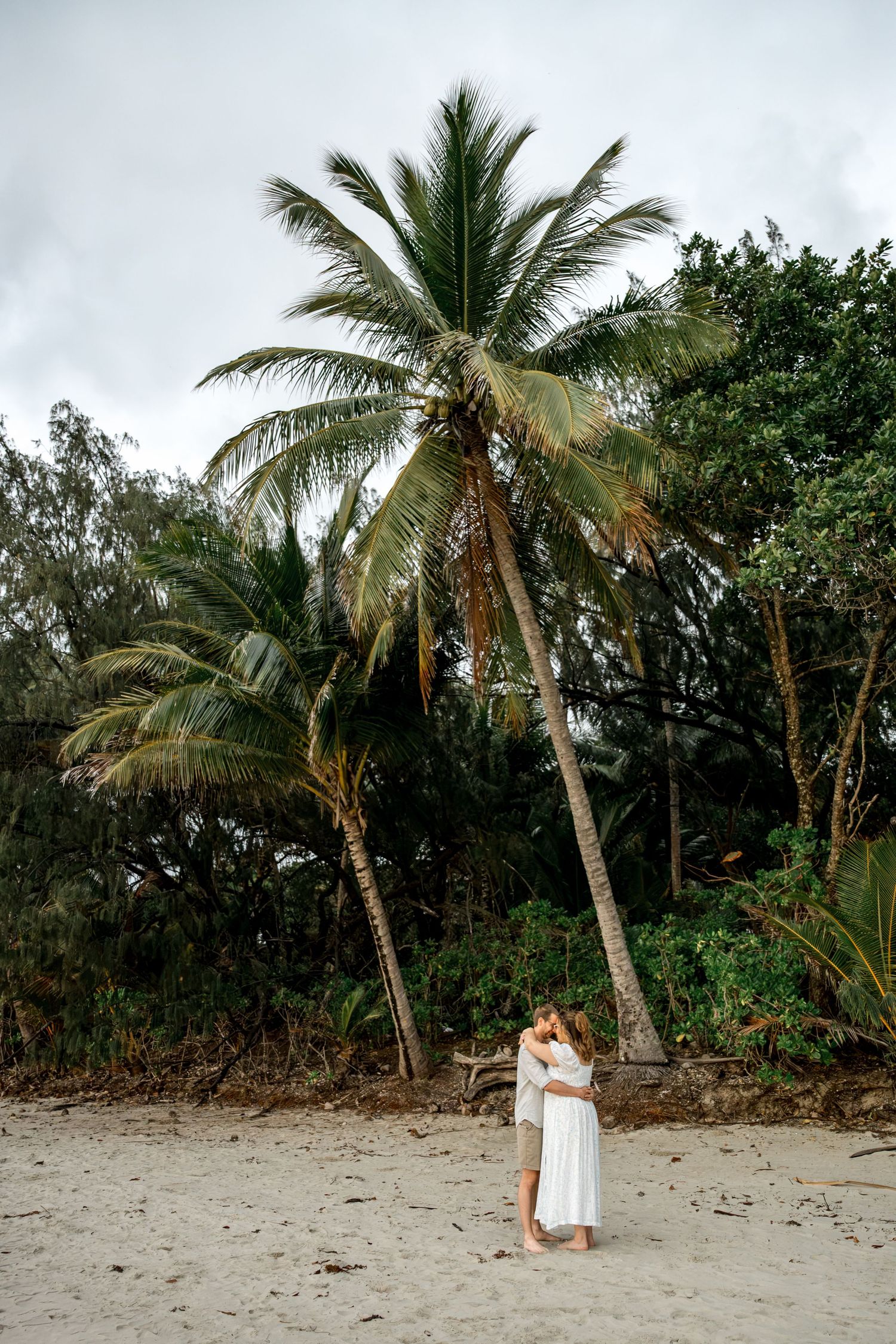 Couple embracing and kissing under tall palm trees on a sandy tropical beach with cloudy sky at Four Mile Beach, Port Douglas.