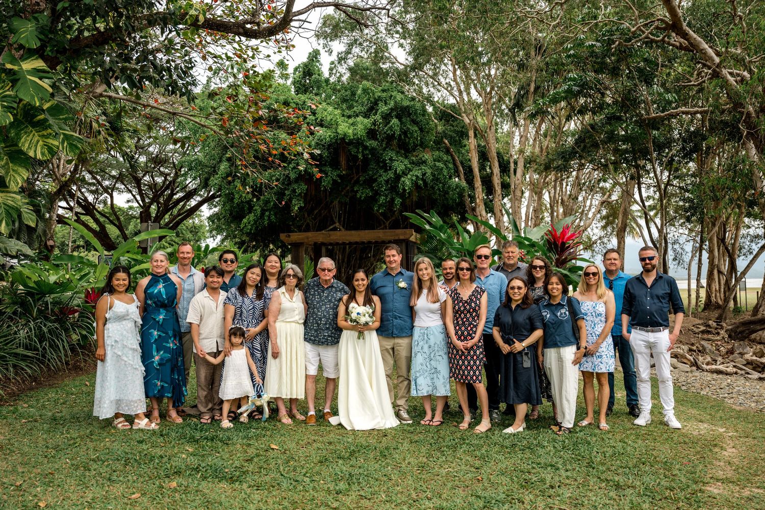 Wedding party and guests pose for a group photo on a tropical garden lawn with lush green trees at Little Cove, Port Douglas.