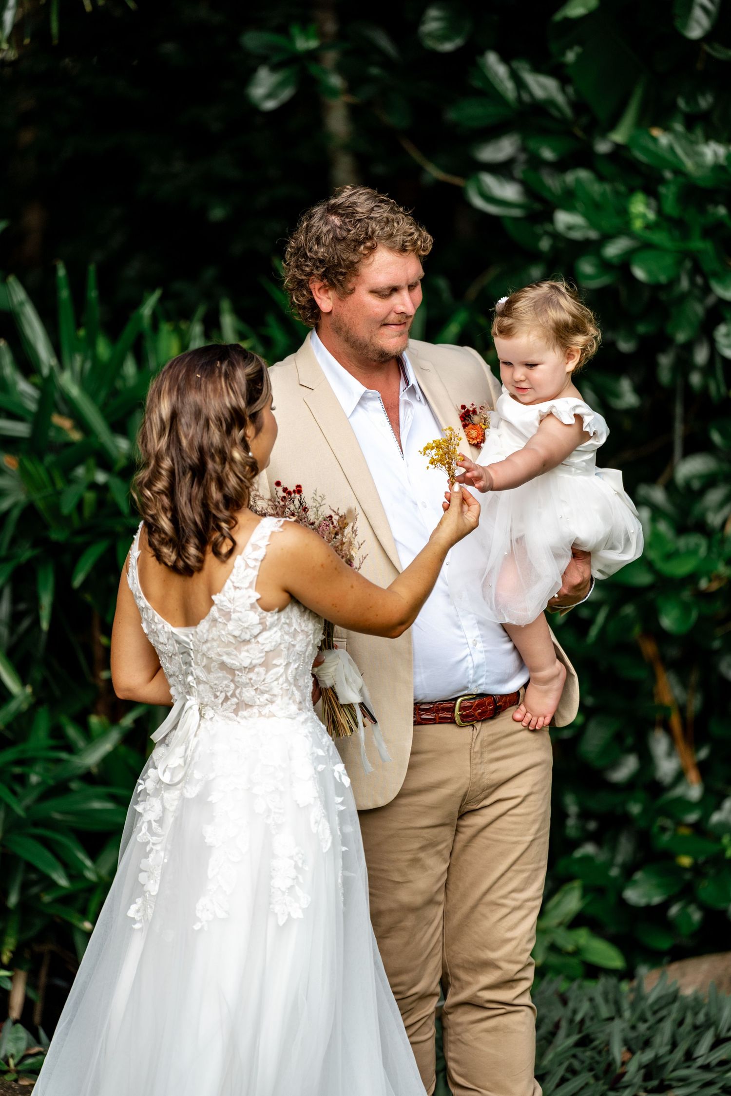 Bride and groom hold their baby daughter during outdoor wedding ceremony in tropical garden.