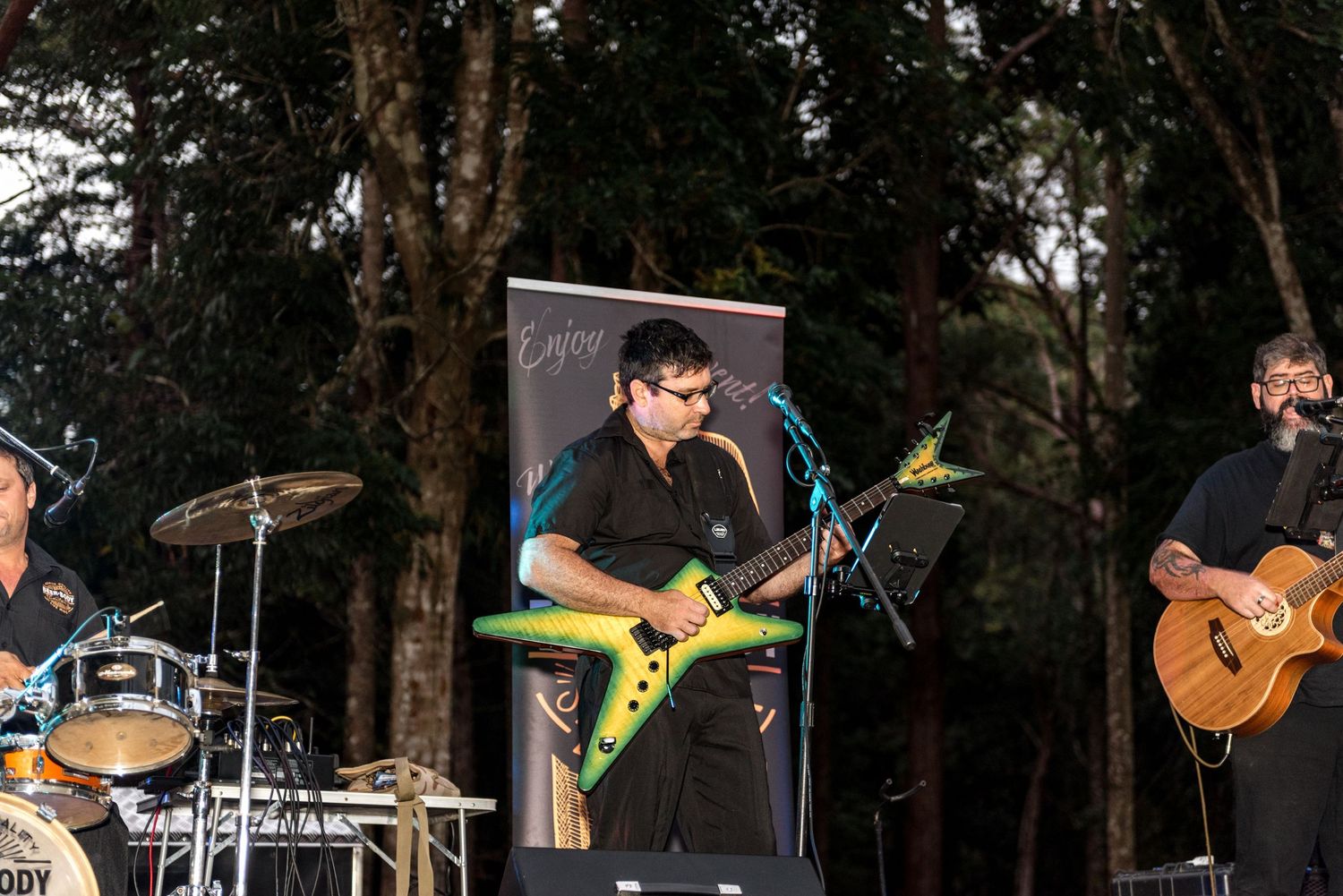 Live band performing outdoors at night with colorful electric guitar under forest tree backdrop.