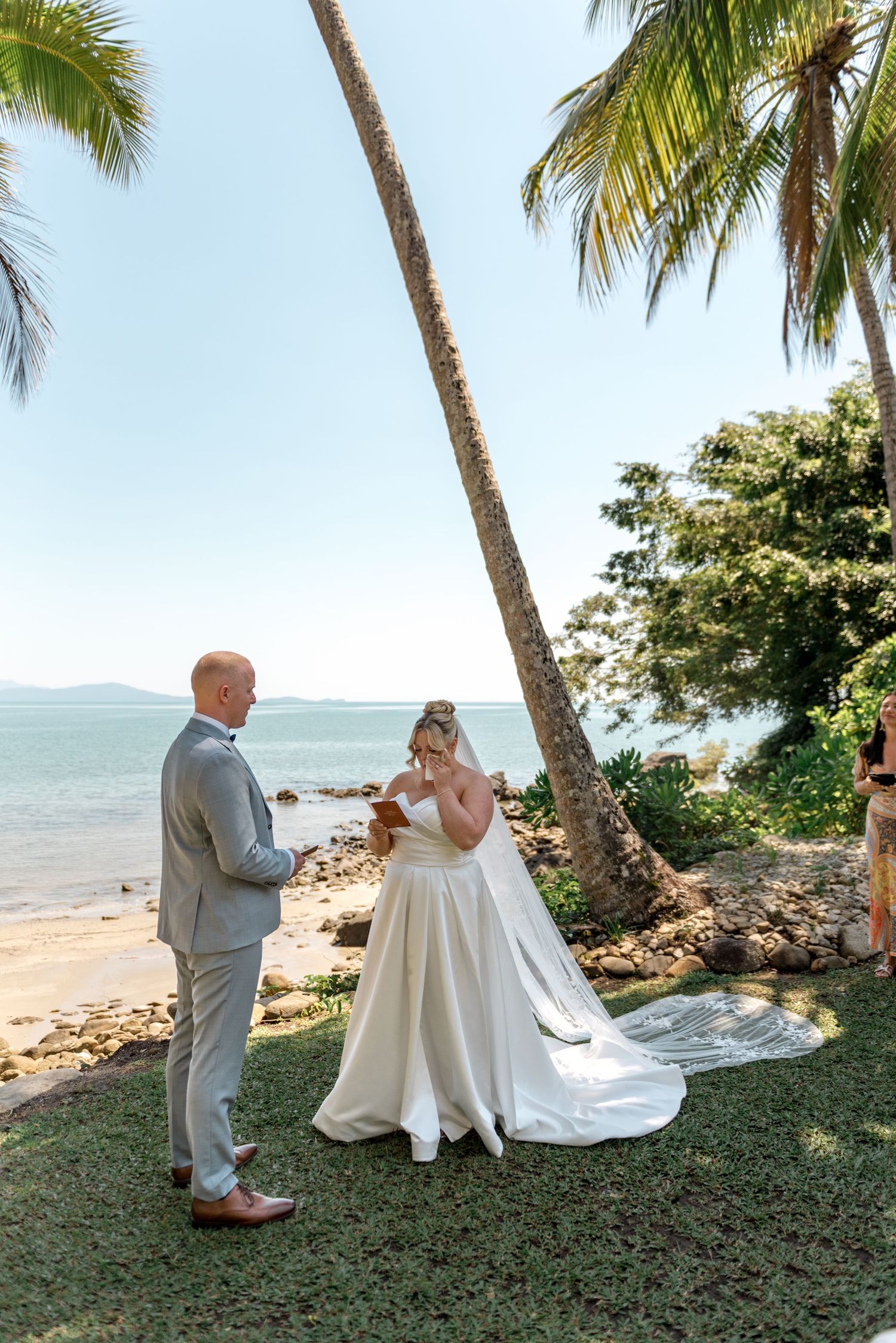 Bride reads vows to groom at tropical beachside wedding ceremony under palm trees.