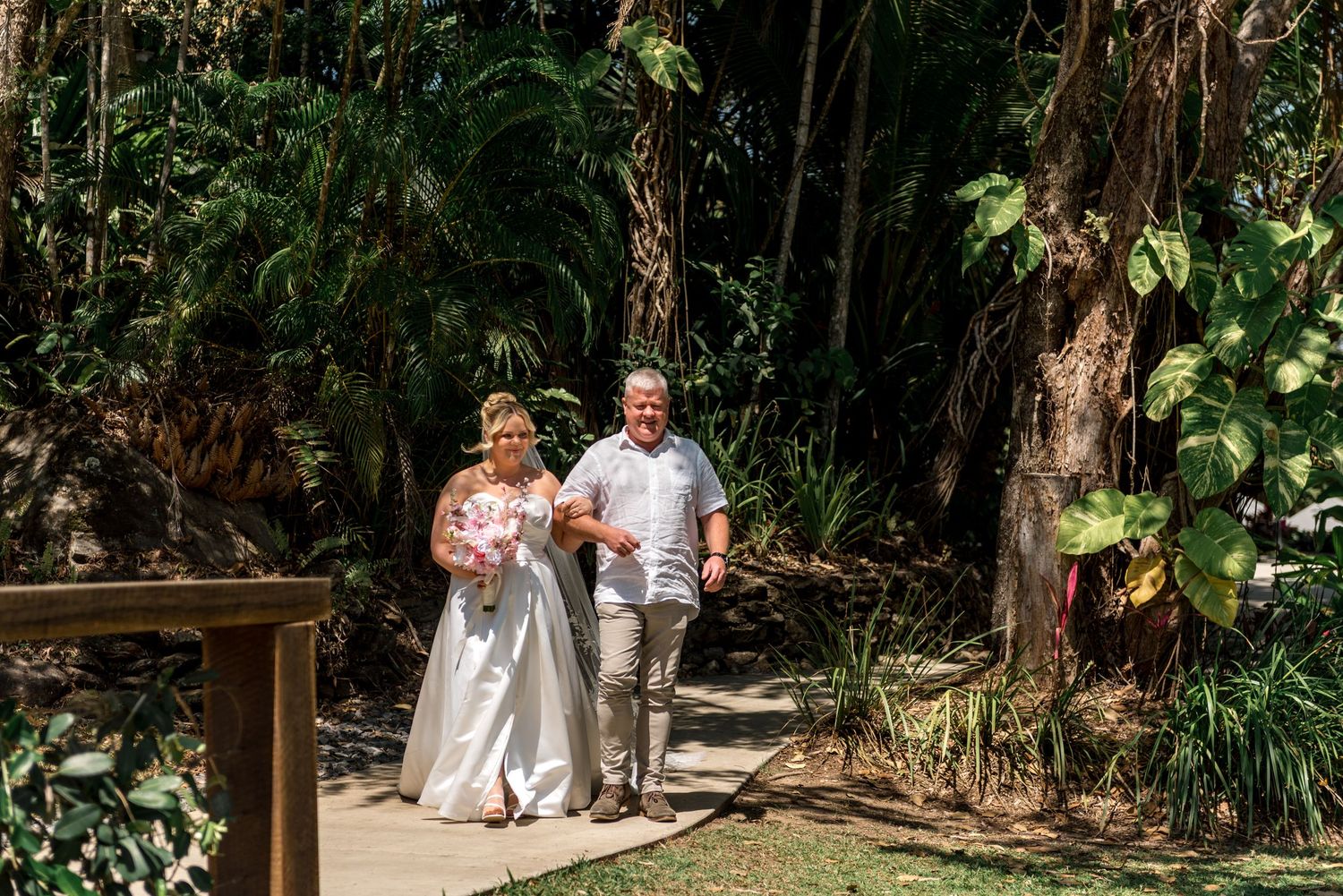 Happy bride and groom walk down tropical garden path during outdoor wedding celebration.