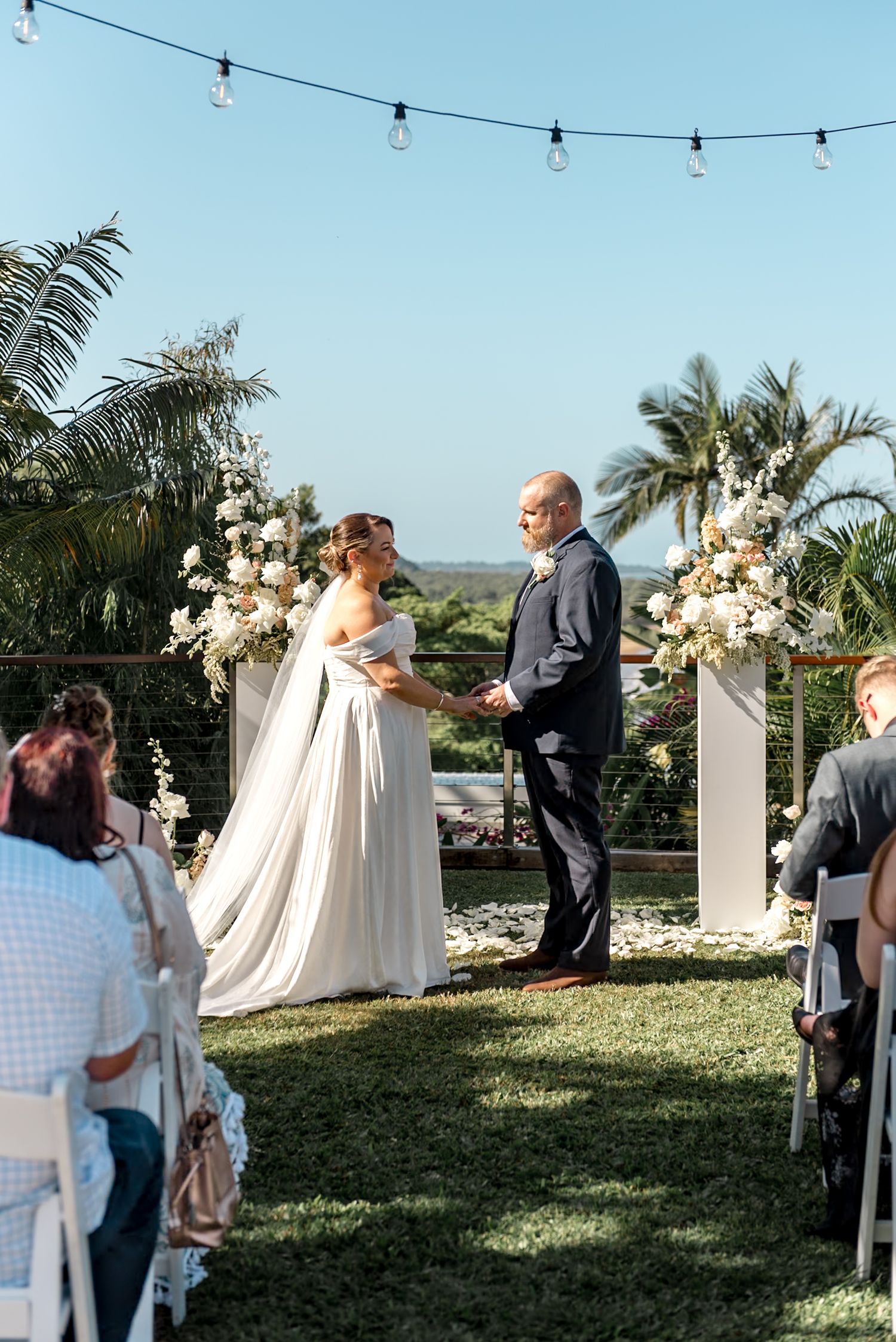 Bride and groom hold hands at outdoor wedding altar adorned with white floral arrangements under blue sky.