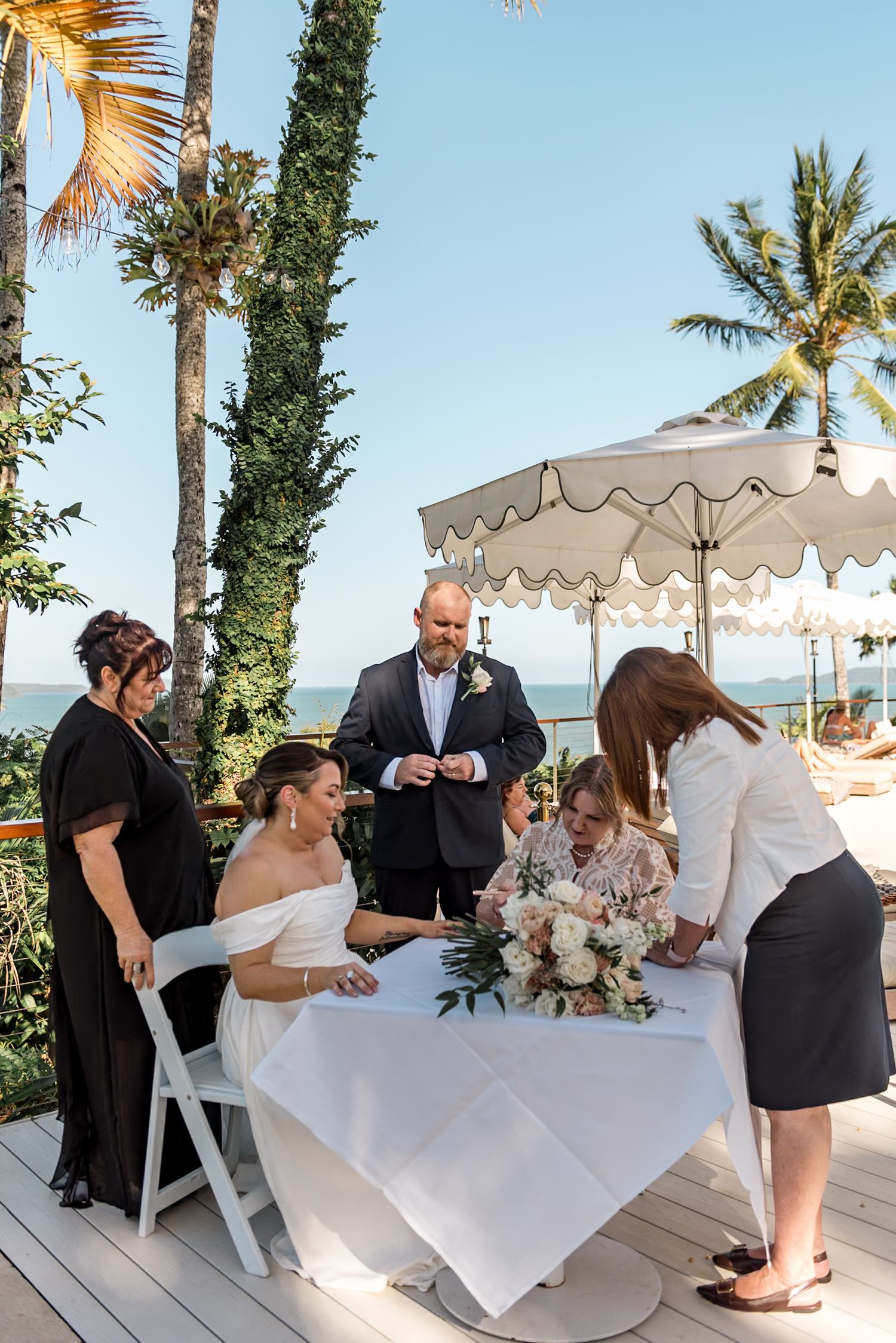 Wedding party gathered around signing table on tropical deck with ocean views and palm trees.