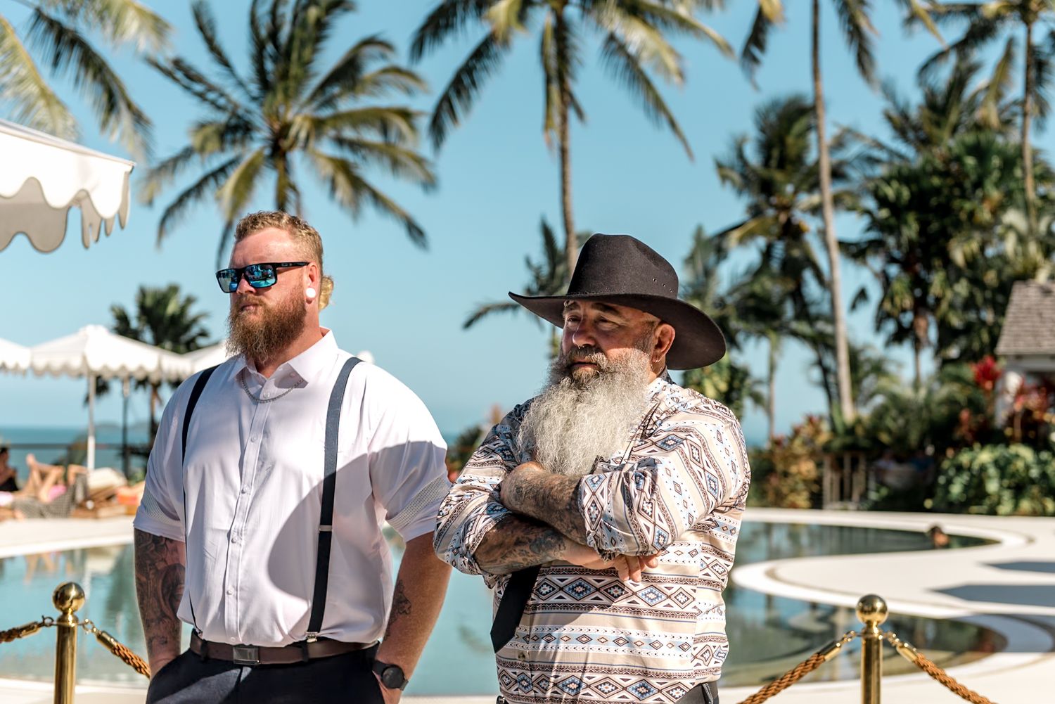 Two male wedding guests stand near pool with palm trees and white umbrellas at tropical venue.