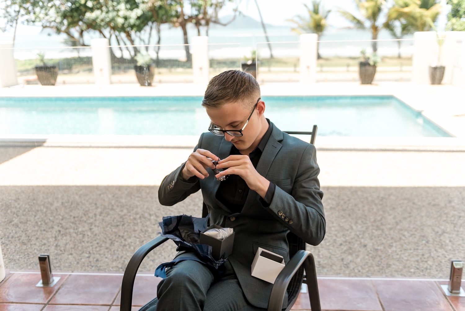 Photographer in dark suit checking camera equipment while seated poolside outdoors.