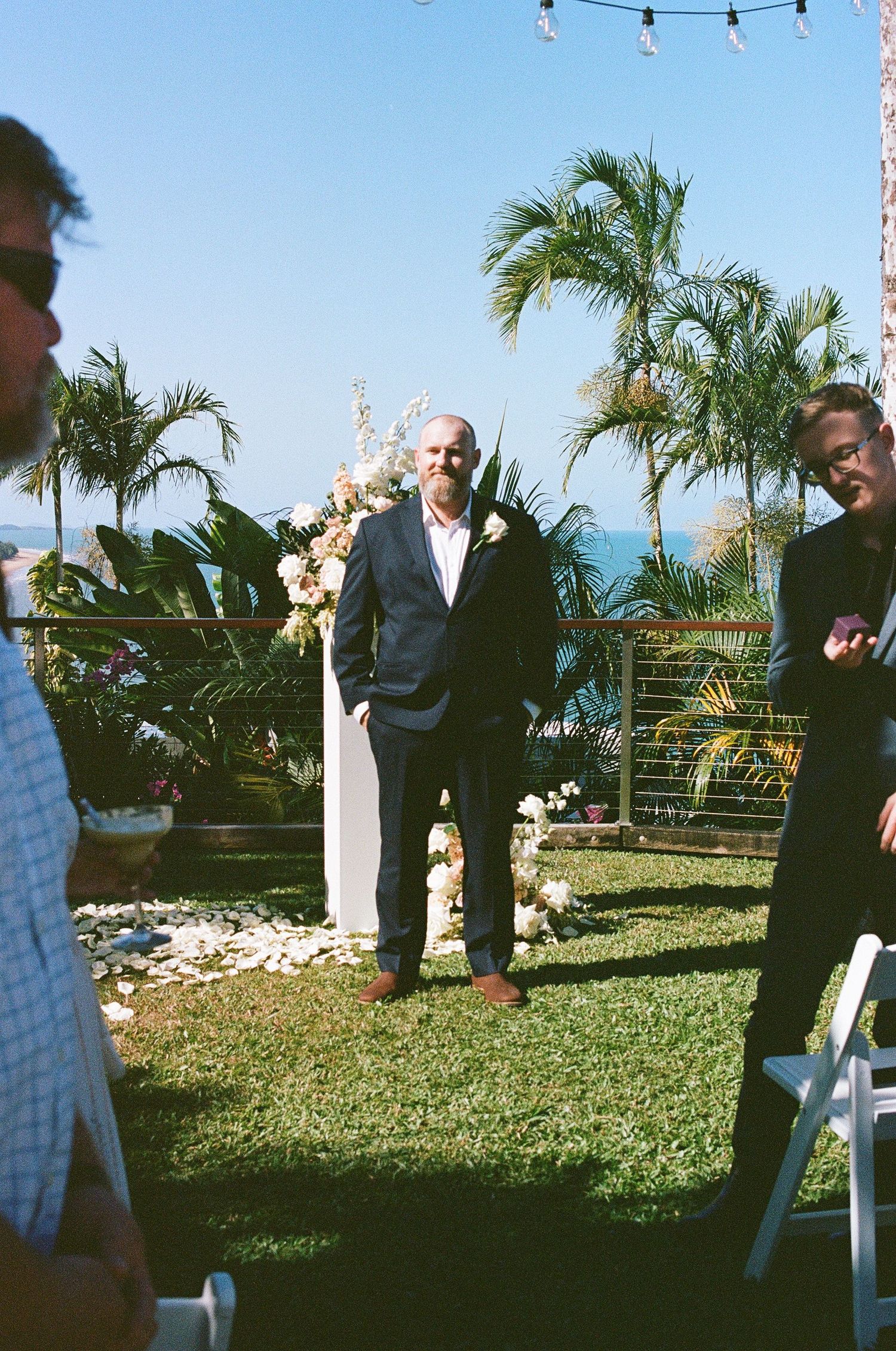 Groom in navy suit standing at floral wedding altar surrounded by palm trees and ocean backdrop.