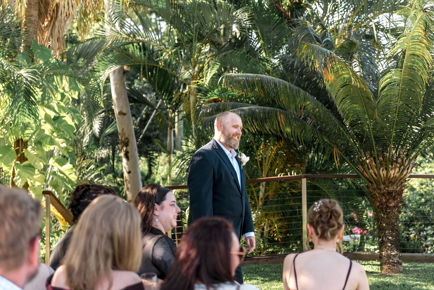 Smiling groom in black tuxedo stands at outdoor wedding ceremony surrounded by tropical greenery.