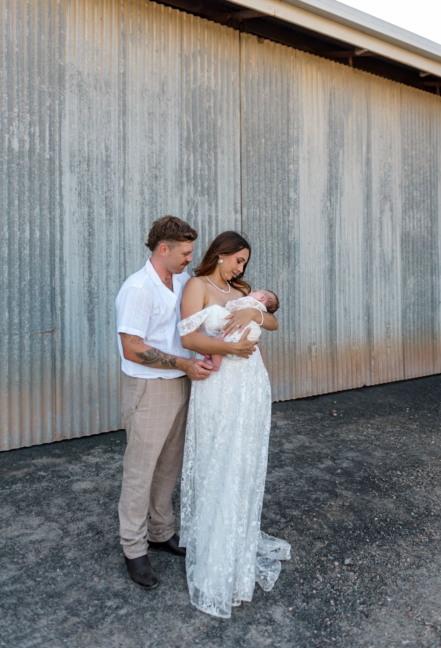 Bride in off-shoulder lace gown and groom in white shirt tenderly holding newborn baby together.