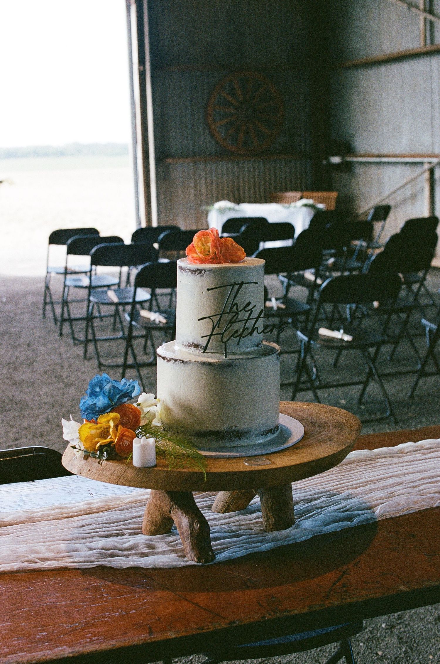 Two-tier white wedding cake with colorful flowers on wooden stand inside a barn venue.
