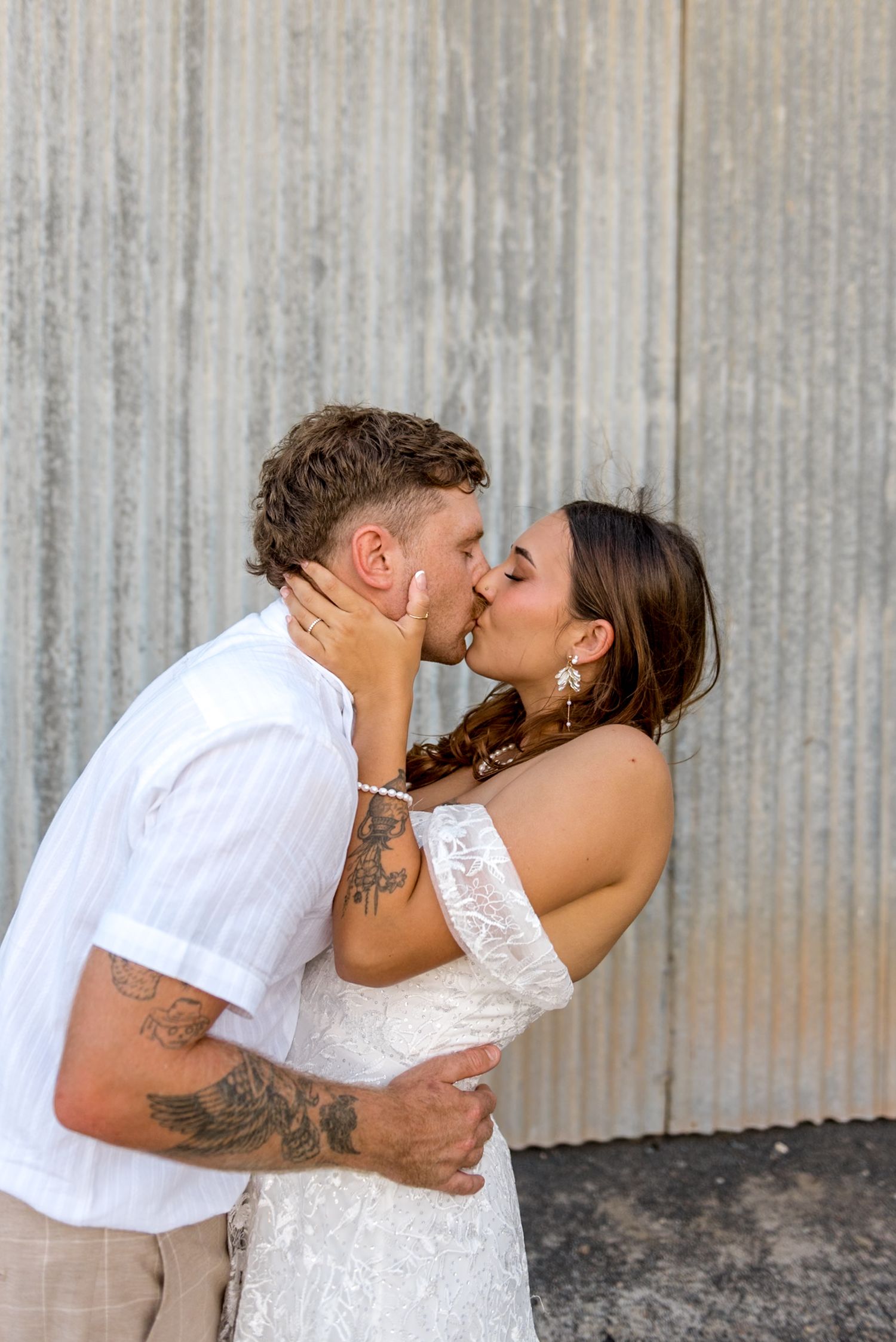 Tattooed groom in white shirt kisses bride in lace off-shoulder gown against corrugated metal wall backdrop.