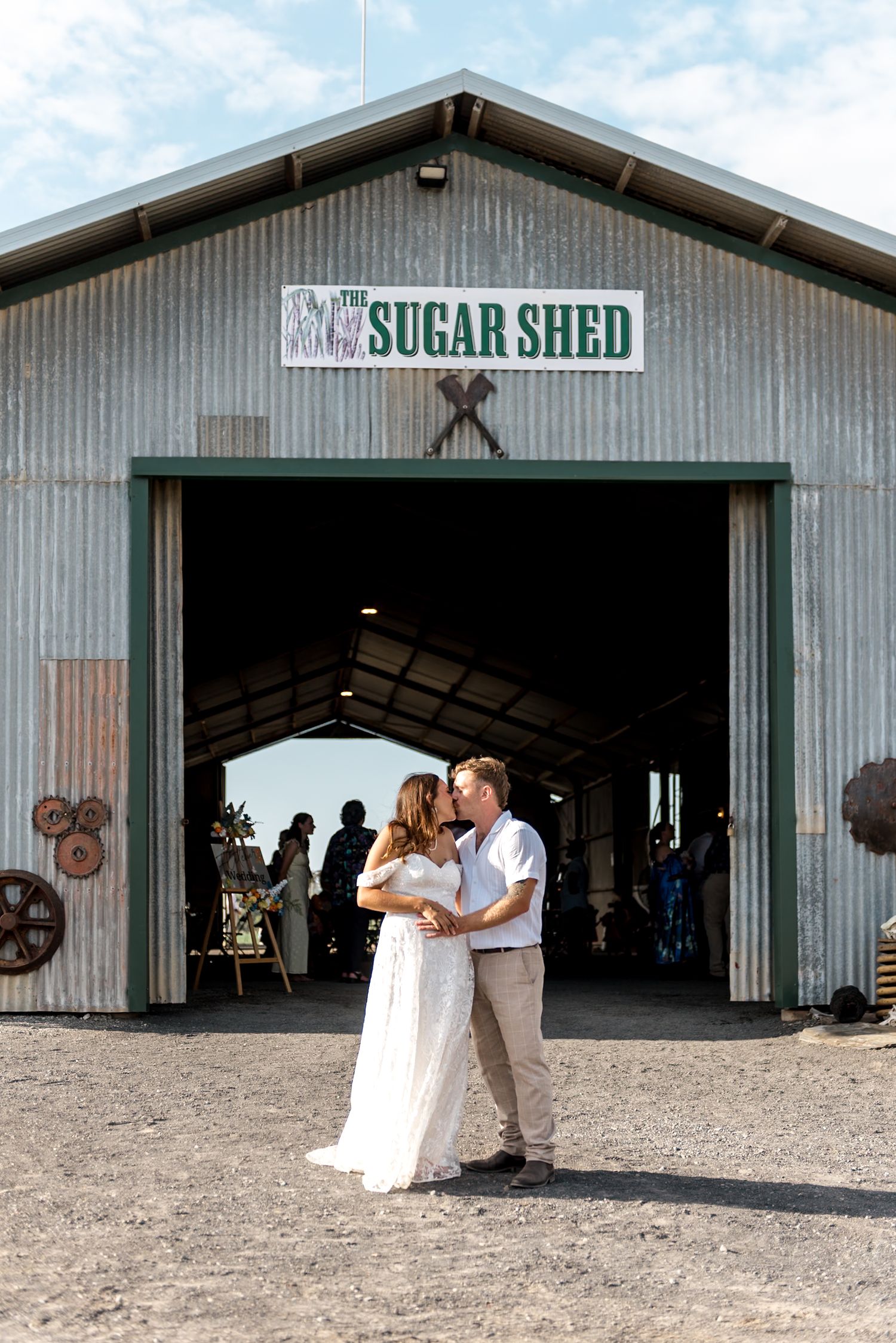 Newlyweds kissing in front of The Sugar Shed wedding venue with guests visible inside.