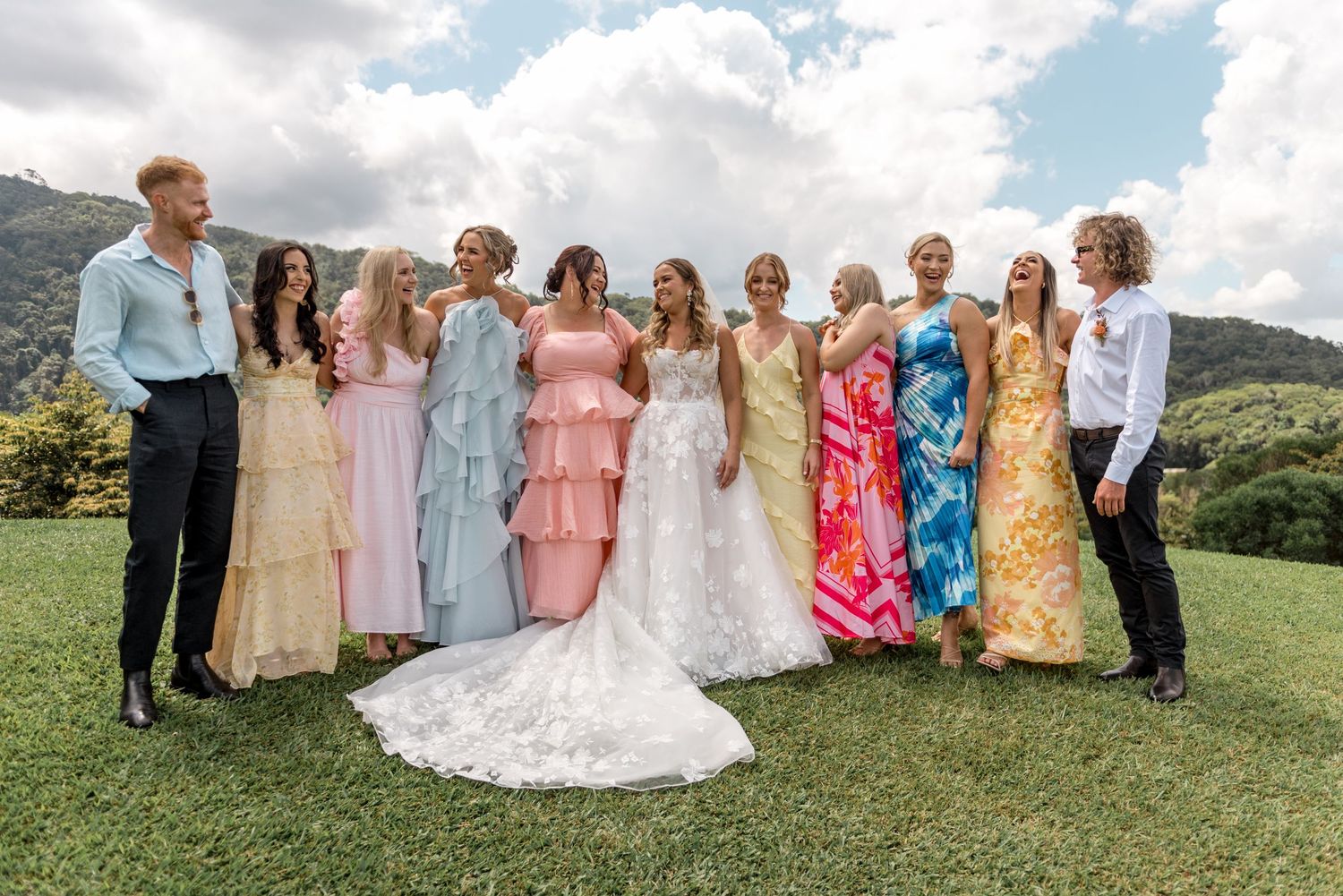 Bride and wedding party in vibrant mixed-color gowns laugh together on green hillside lawn.