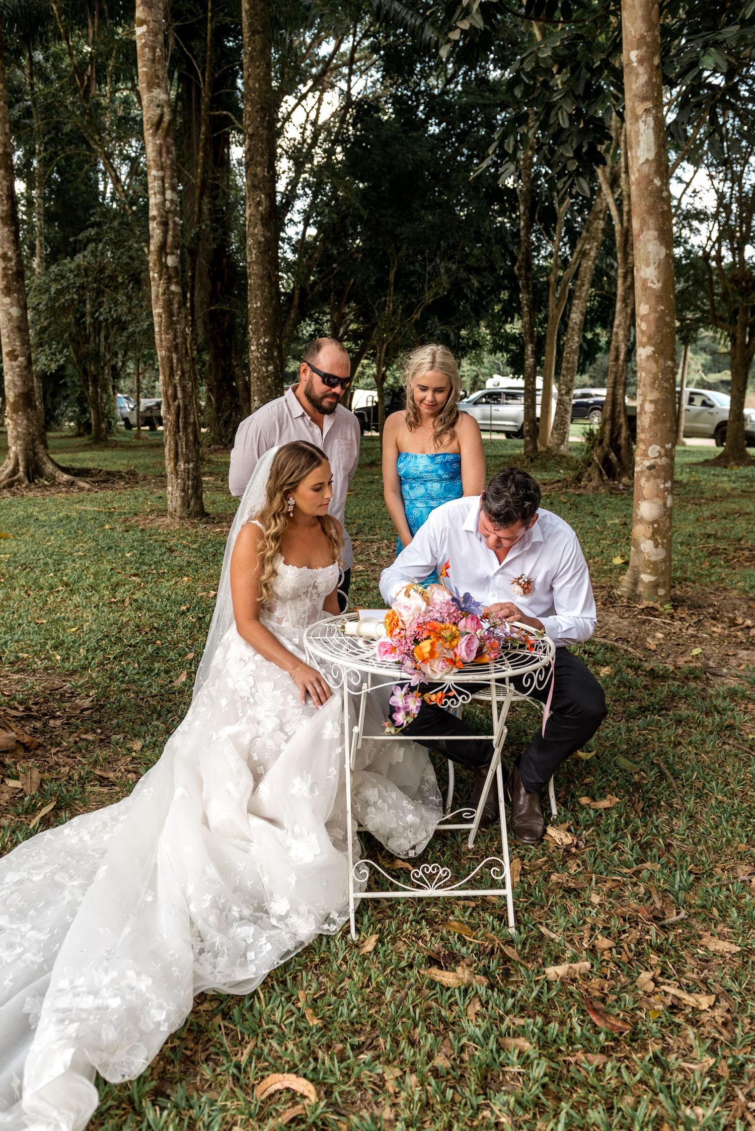 Bride and groom sign wedding documents at an ornate white table surrounded by witnesses in a lush forest setting.
