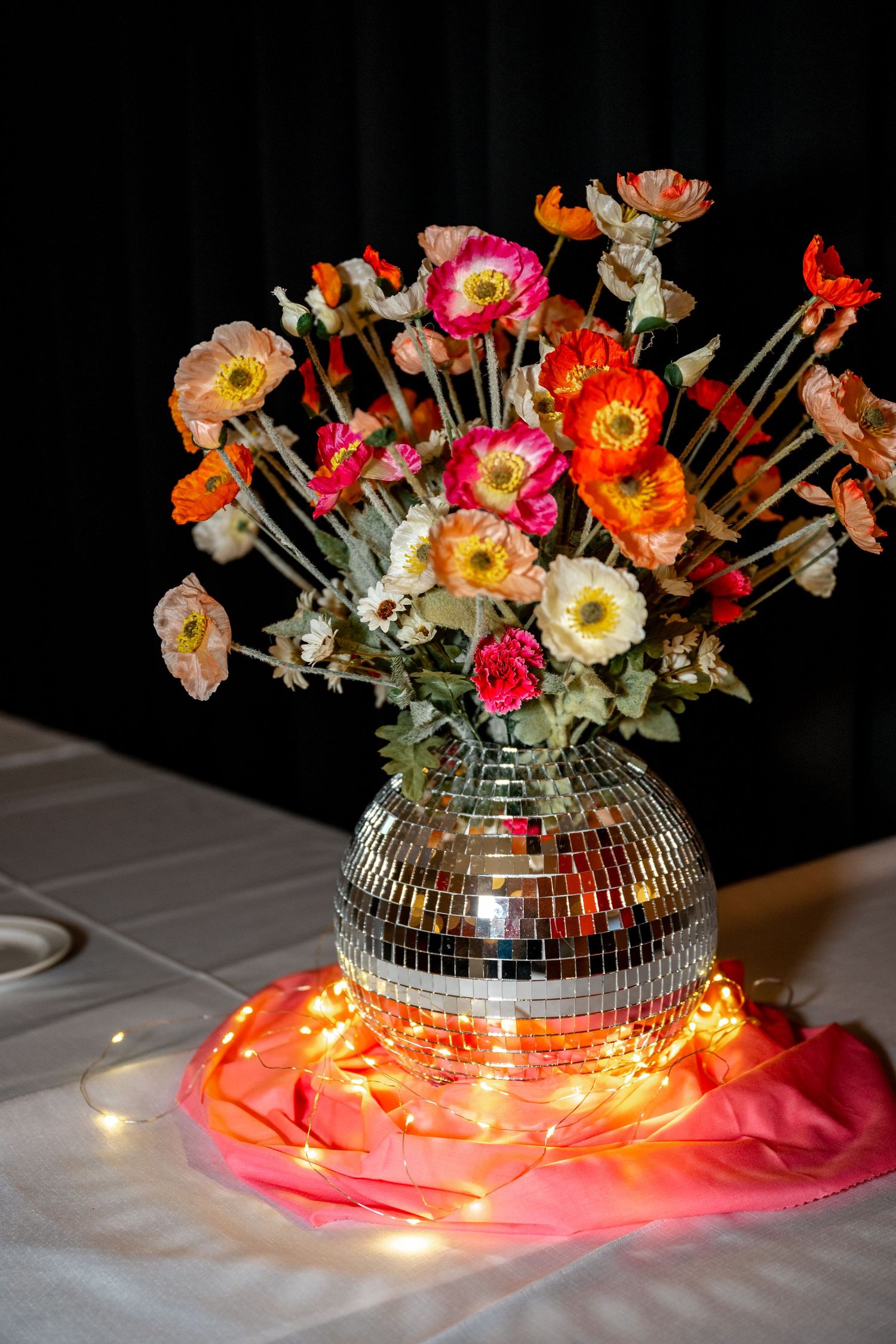 Colorful poppies arranged in a mirrored disco ball vase with warm fairy lights on a white tablecloth.