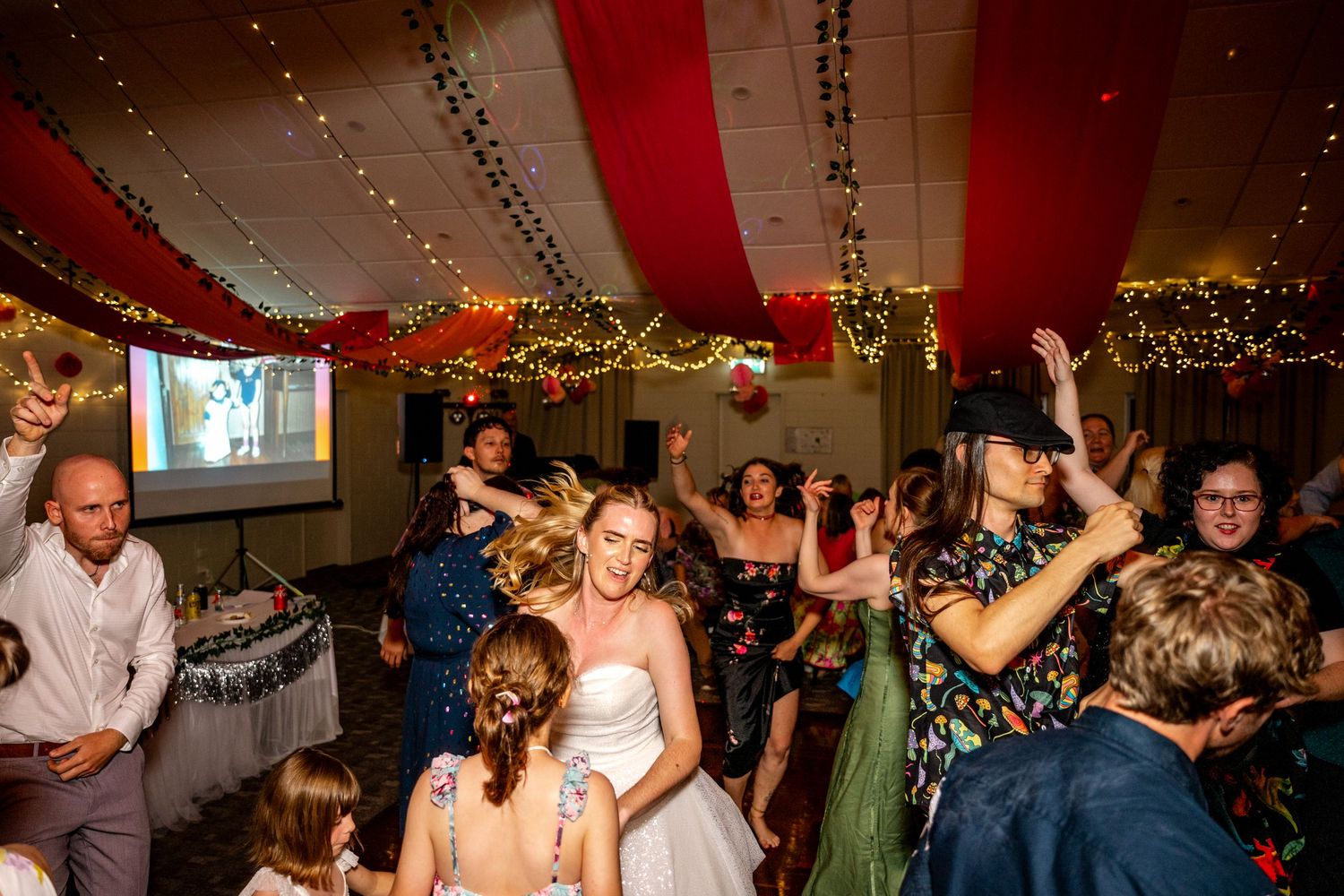 Bride dancing joyfully with wedding guests under fairy lights at reception.