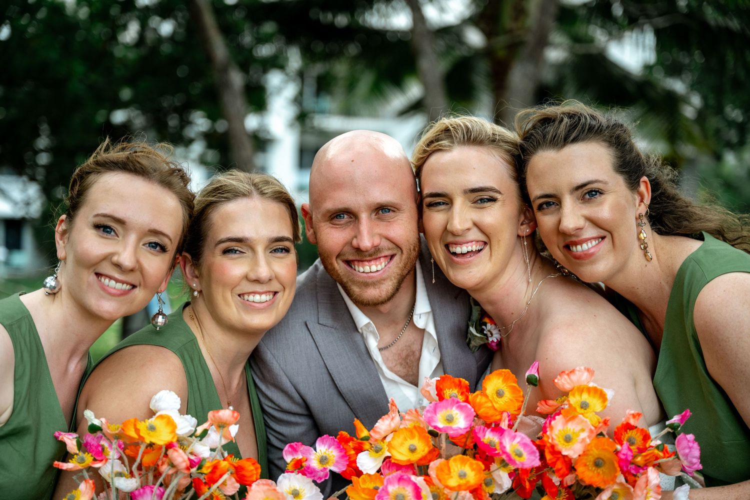Groom in grey suit smiles surrounded by four bridesmaids in green dresses holding bright orange and pink bouquets.