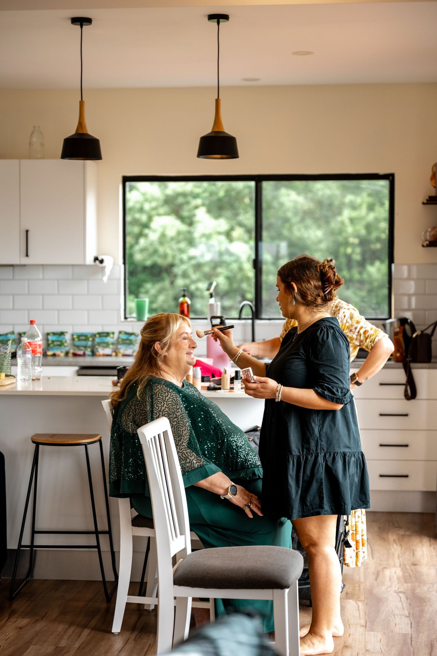 Wedding makeup artist applying makeup to seated woman in green sequin top in modern kitchen.