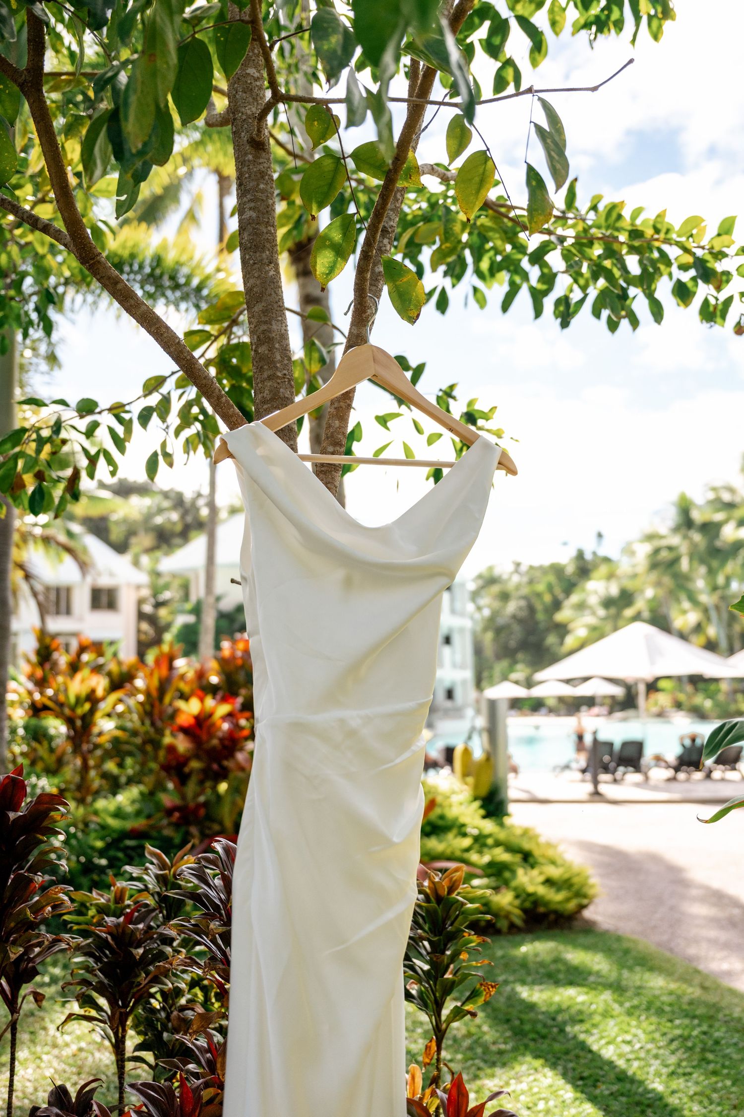 Simple white wedding dress hanging on wooden hanger from tree branch in tropical resort garden setting.