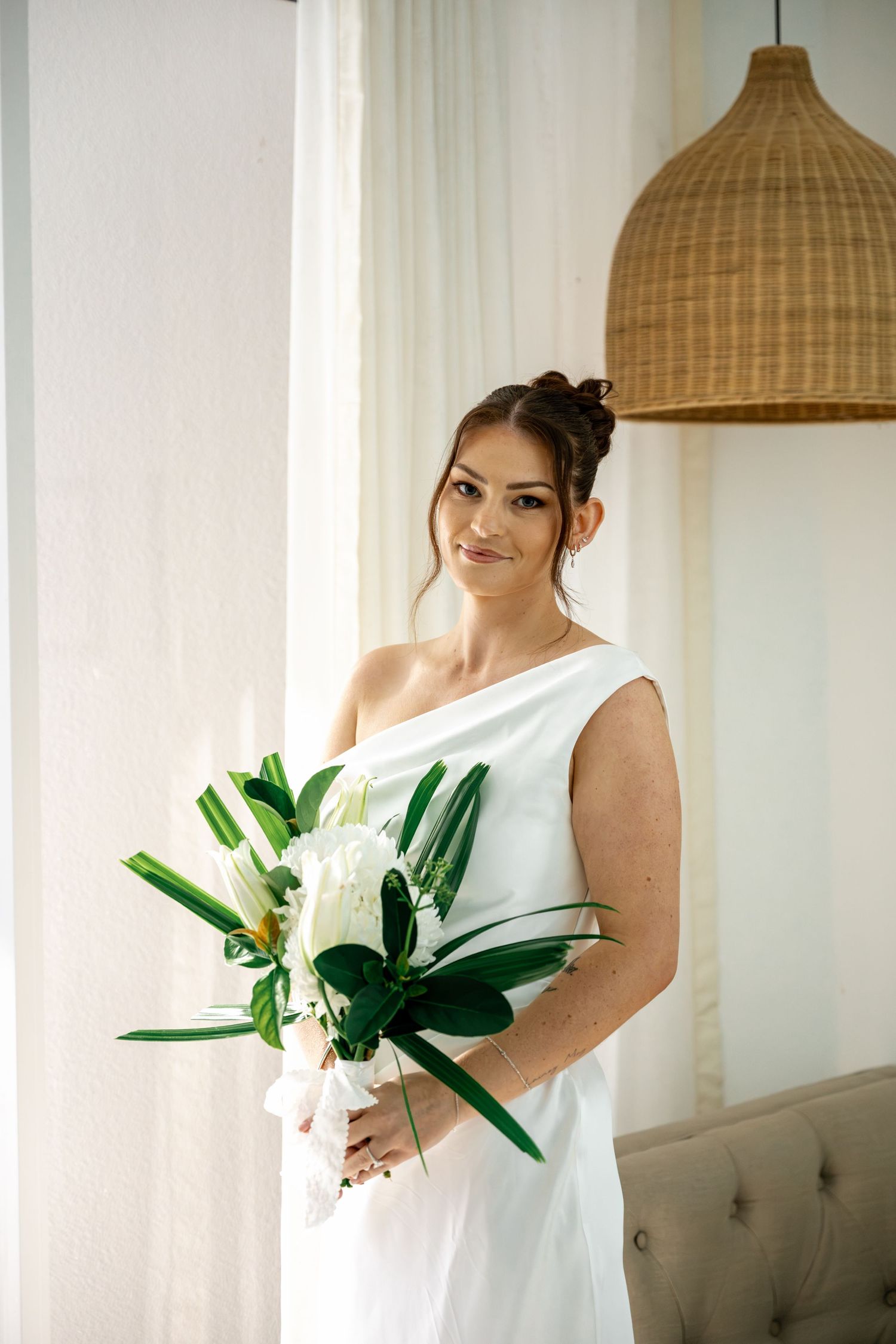 Elegant bride in white one-shoulder gown holding tropical bouquet with green leaves and white flowers indoors at Sheraton Mirage Resort.