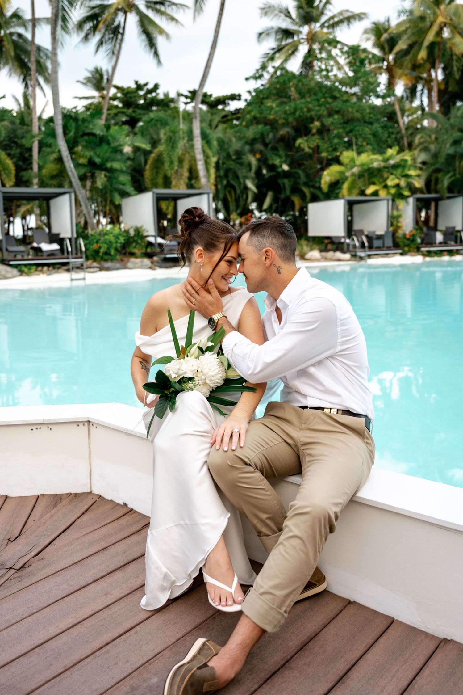 Bride and groom sitting poolside at a tropical resort, sharing an intimate moment with a white bouquet at Sheraton Grand Mirage Port Dougla.
