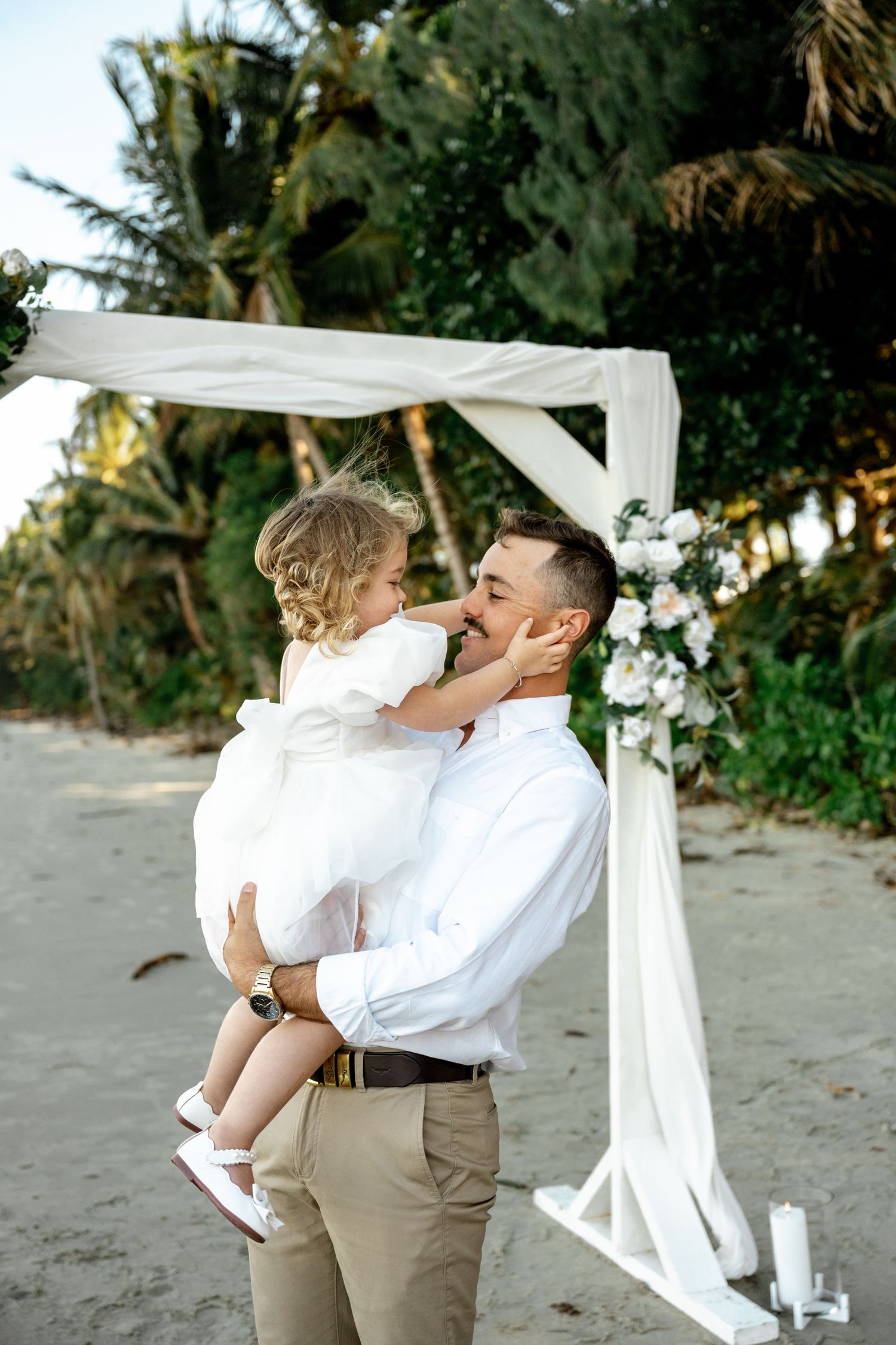 Man in white shirt and toddler girl in white dress share a tender moment at a beach wedding arch at Four Mile Beach, Port Douglas.