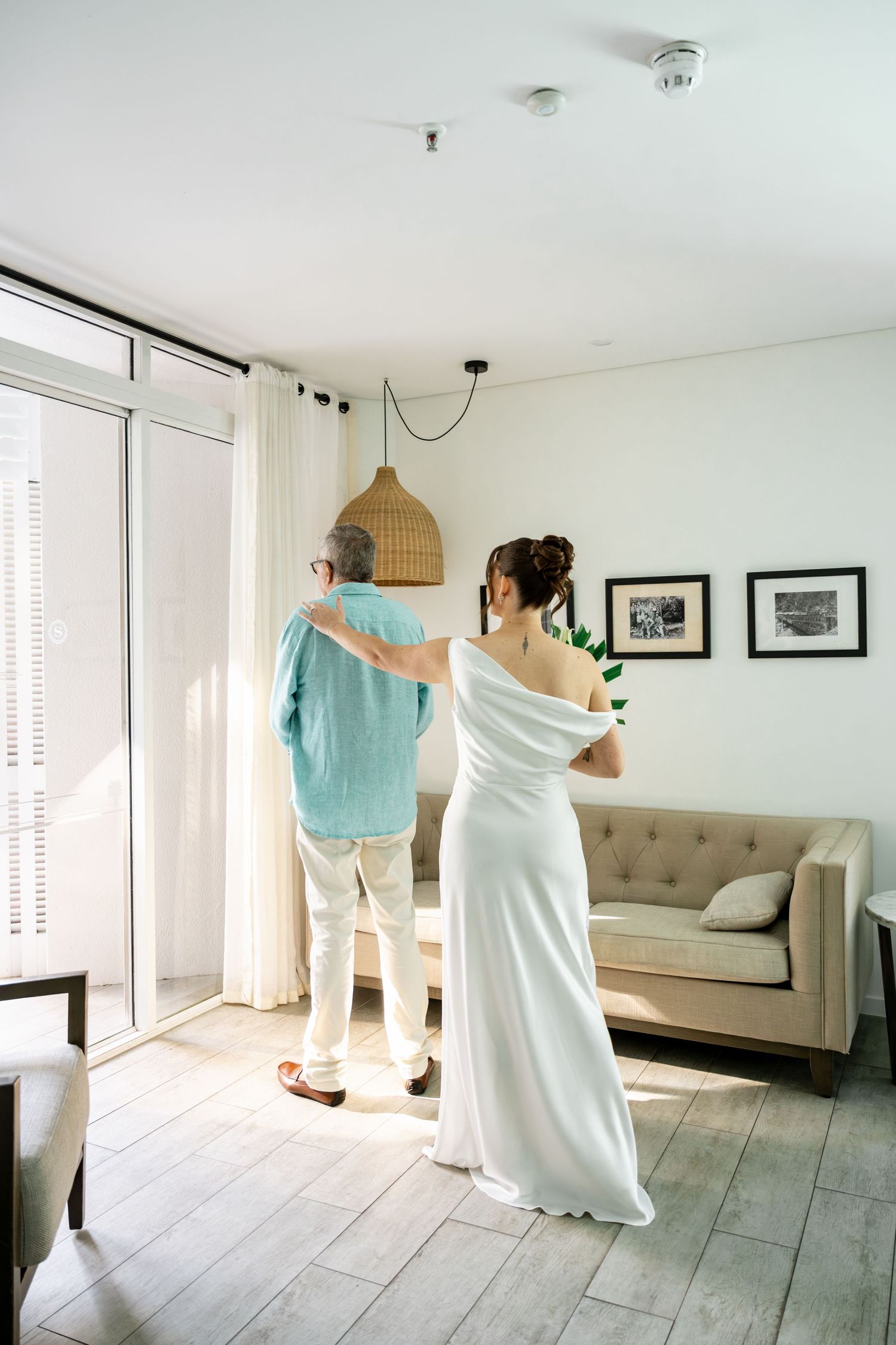Bride in white gown placing hand on older mans shoulder near bright glass doors in modern white room at Sheraton Grand Mirage Resort.