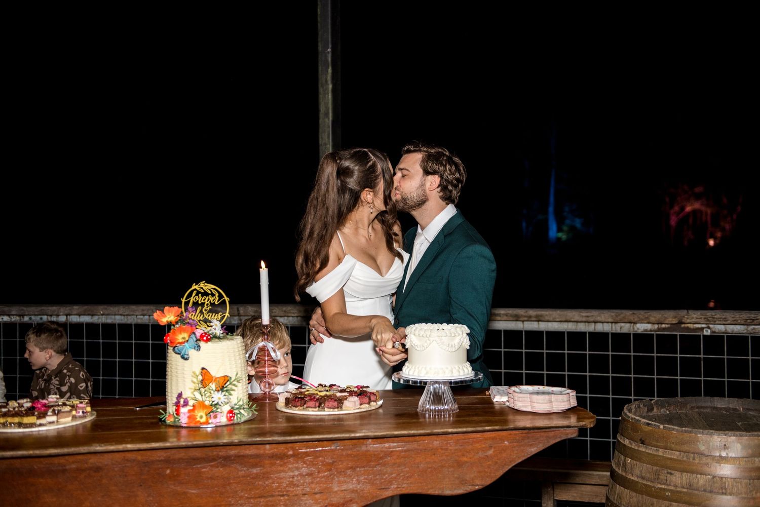 Bride and groom share a romantic kiss at their wedding cake table at night.