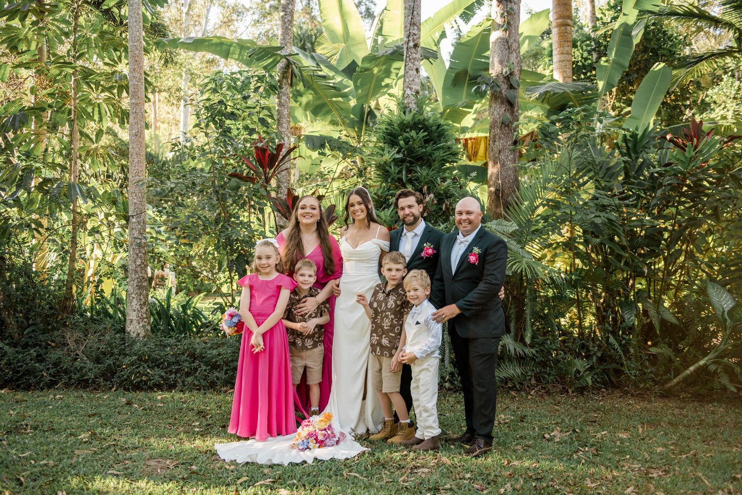 Bride and groom pose with family including children in pink dresses in lush tropical garden.