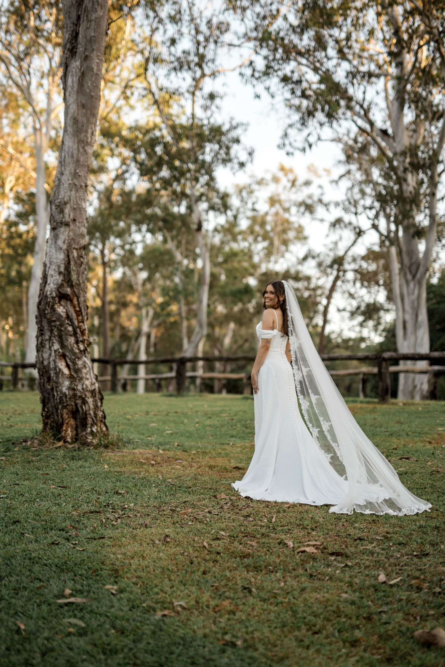 Bride in elegant off-shoulder gown with long lace veil standing in a scenic garden.