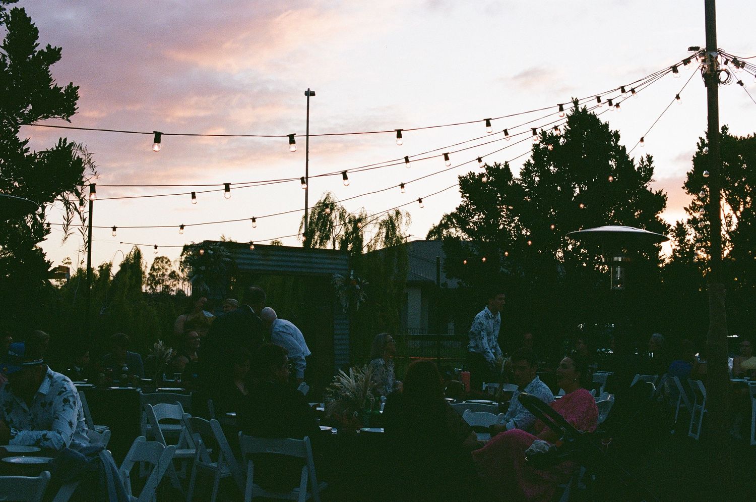 Outdoor wedding reception at dusk with string lights overhead and guests seated at tables under a colorful sky.
