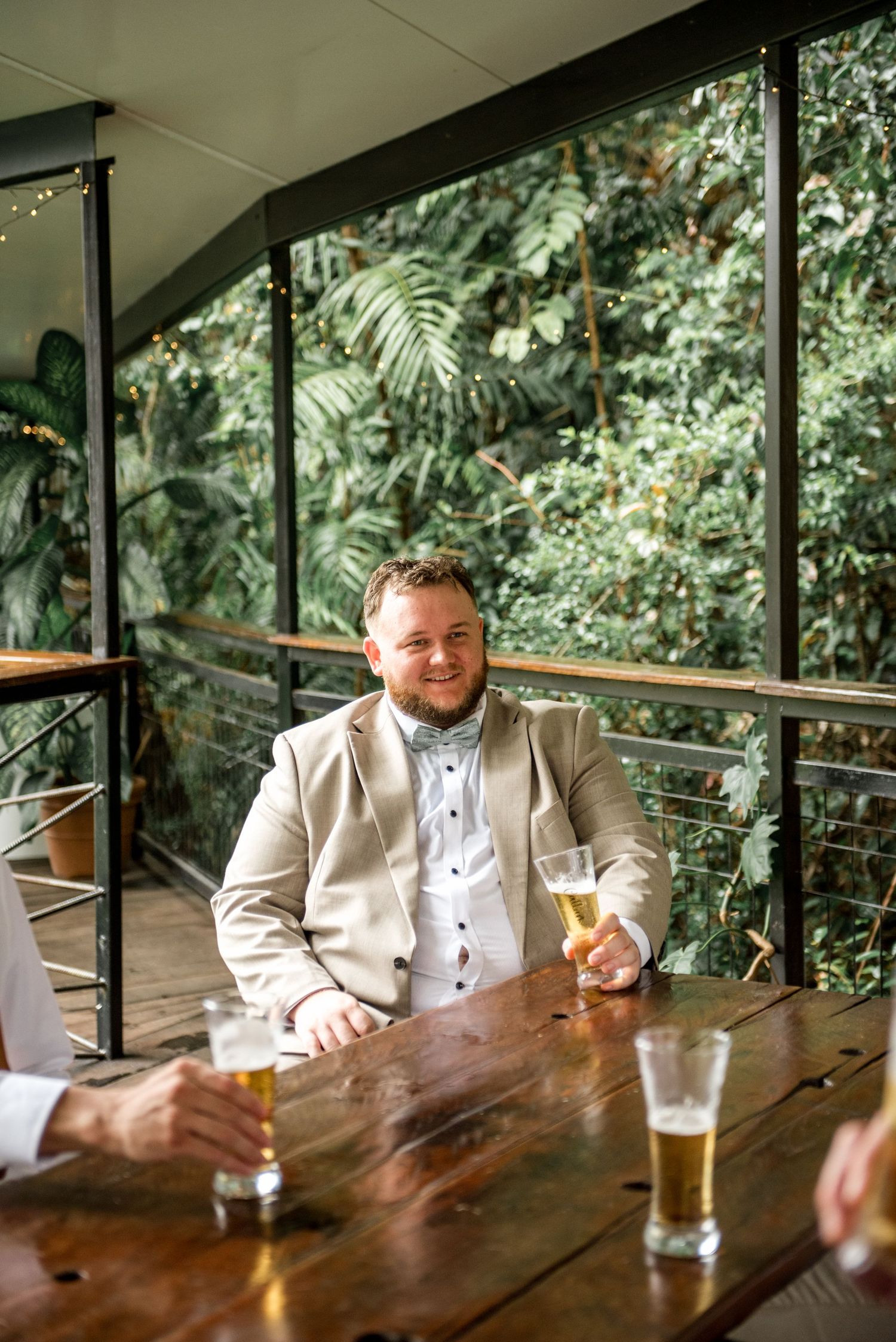 Smiling groom in beige suit enjoying beer with groomsmen on outdoor deck surrounded by greenery.