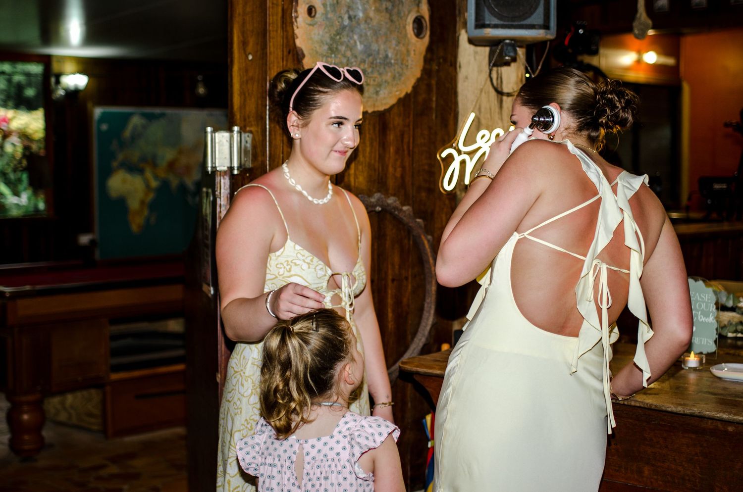 Two bridesmaids in cream dresses chat with a young flower girl inside rustic wedding venue.