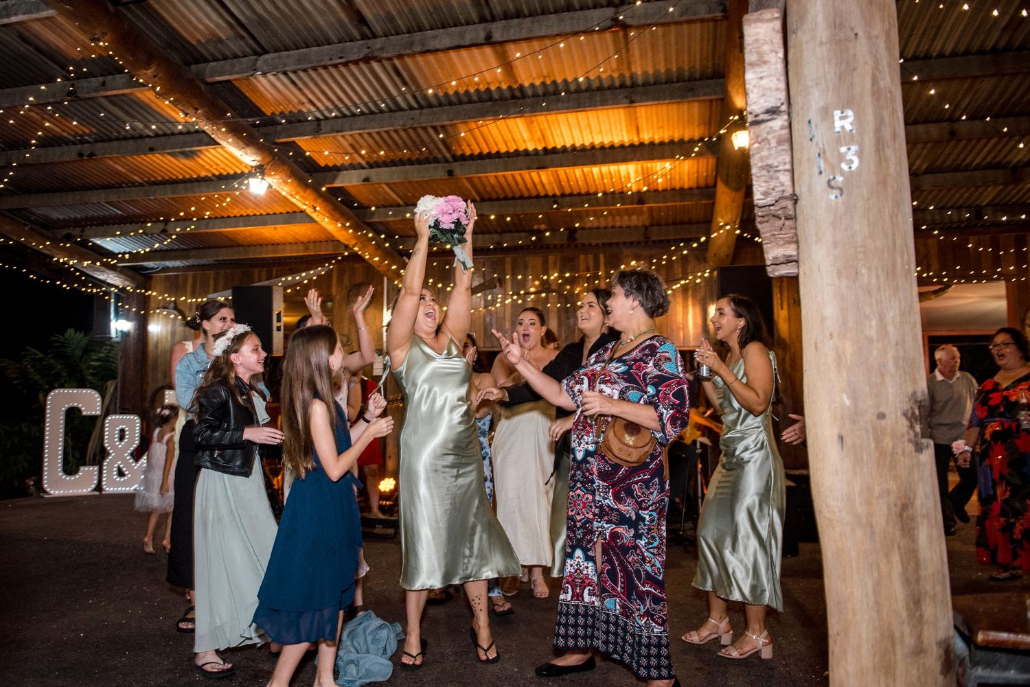 Guests reach for a bouquet toss at a rustic wedding reception decorated with string lights.