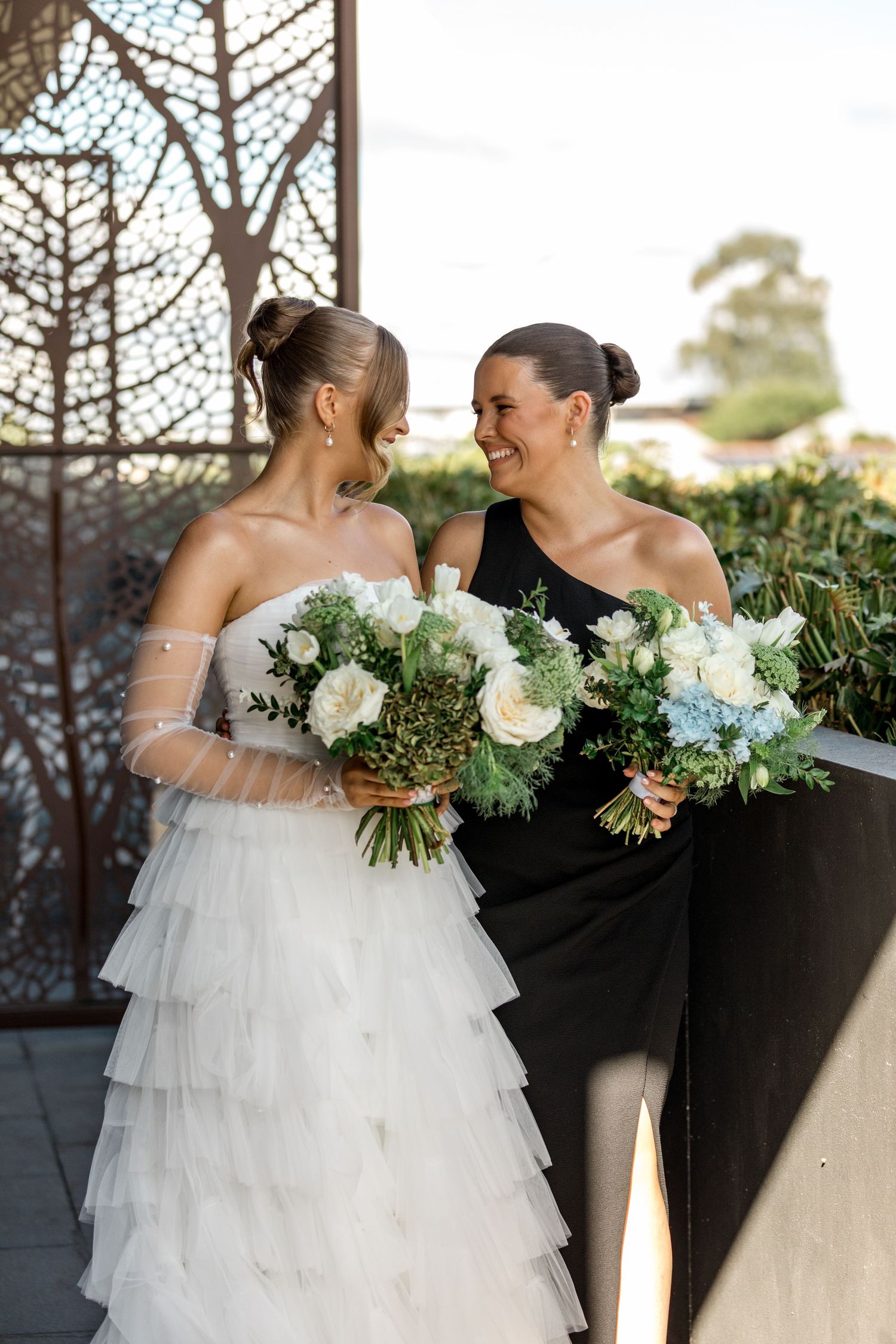 Smiling bride and bridesmaid in black one-shoulder dress hold bouquets on decorated venue balcony.