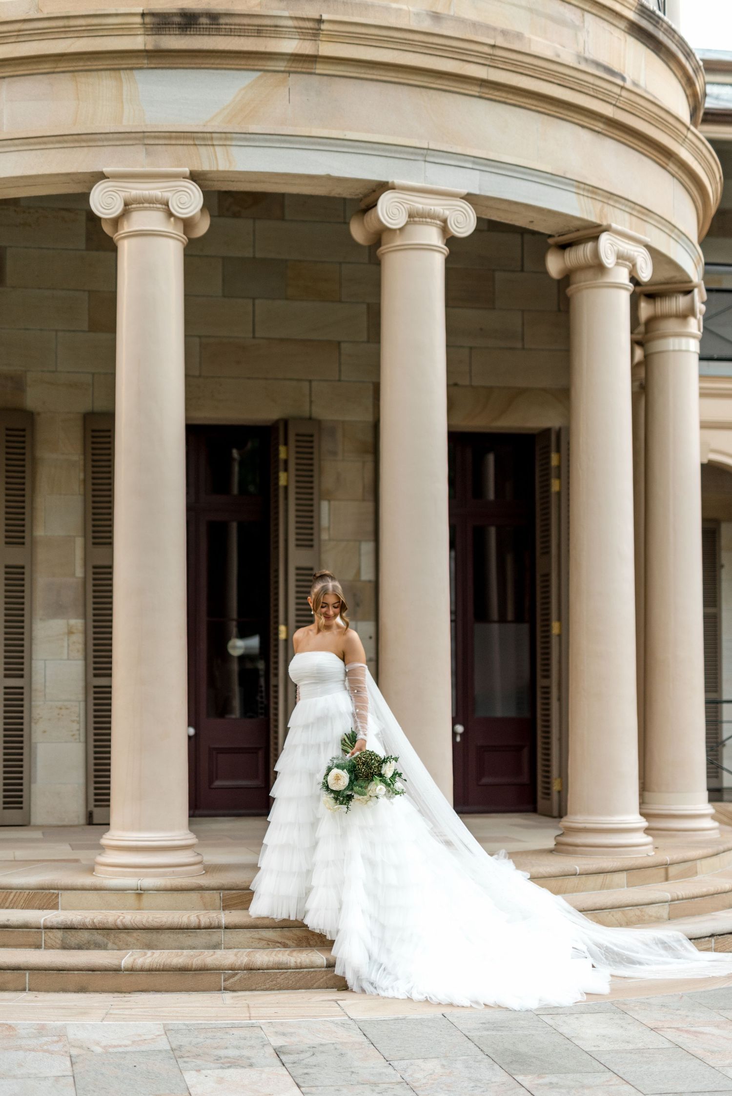Bride in strapless tiered tulle gown holding bouquet before grand colonial sandstone building.