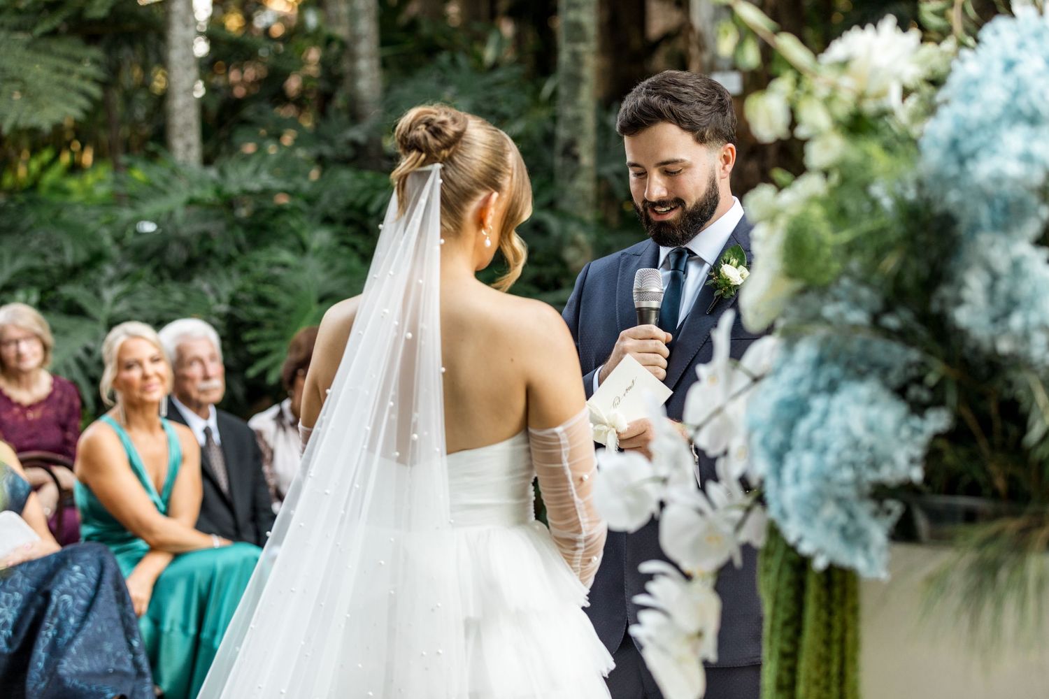 Groom in navy suit smiles at bride while holding microphone during outdoor garden wedding ceremony.